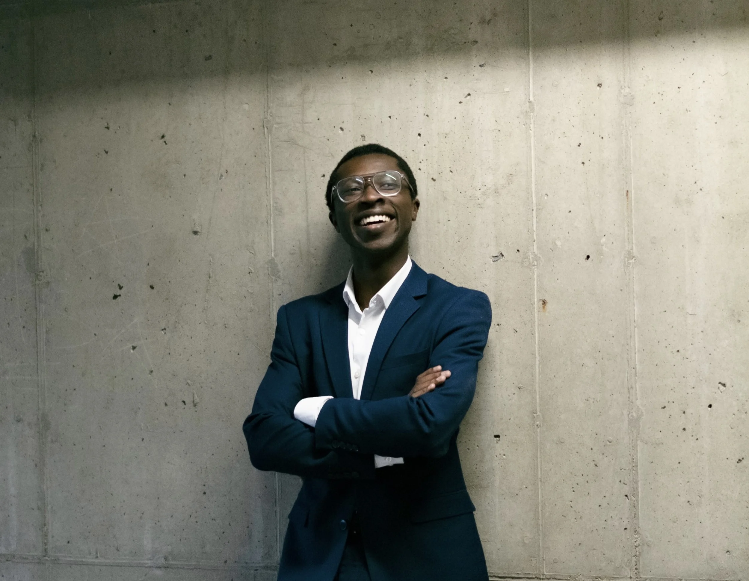 A smiling man in glasses wearing a navy blue suit and white shirt stands against a concrete wall with his arms crossed.