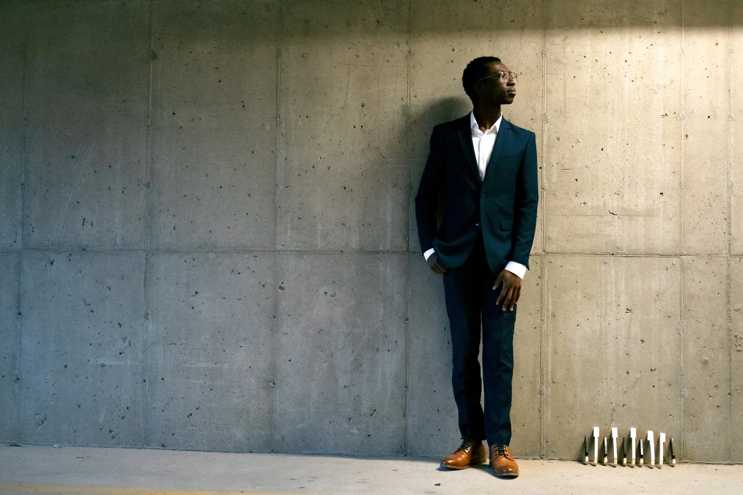 A man in a dark suit with white shirt and brown shoes standing against a concrete wall, looking to his right.