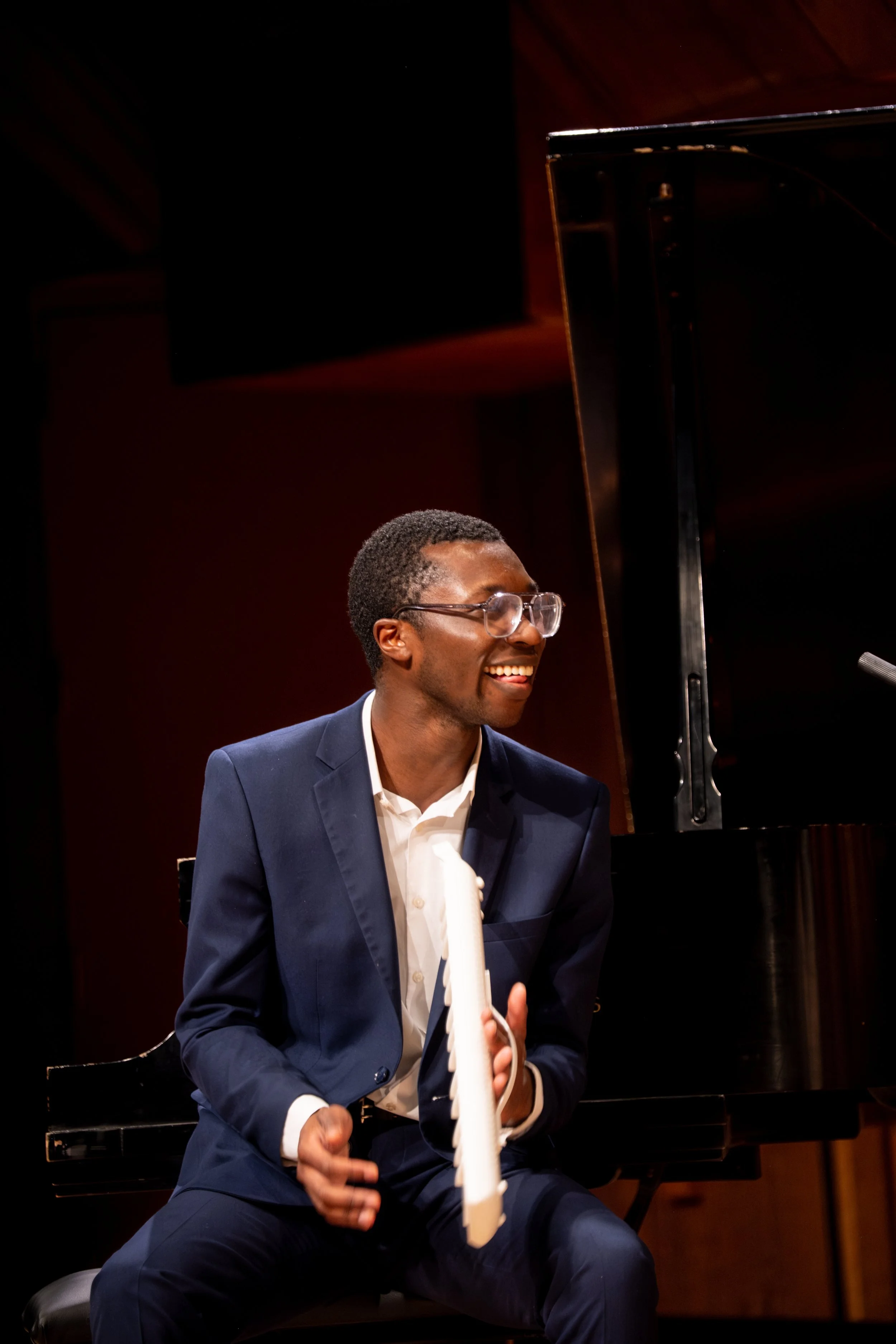 A young man with glasses, wearing a blue suit, is sitting at a piano, smiling and holding a white object in his right hand, in a dimly lit room.