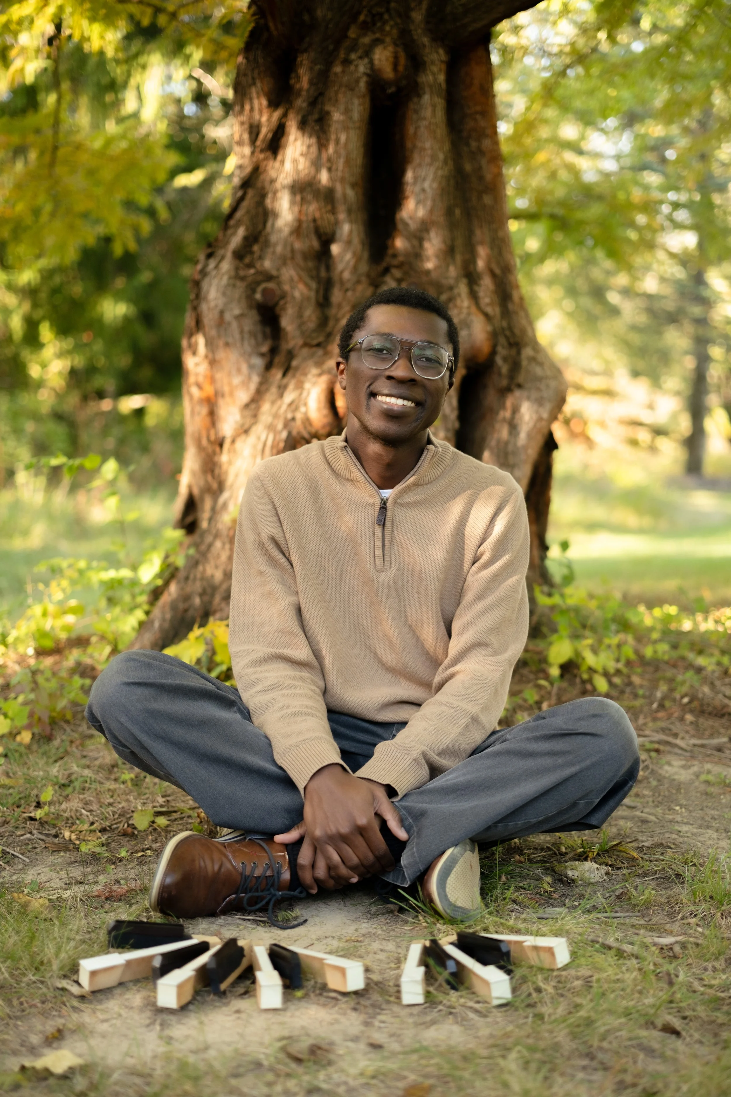 A young man sitting cross-legged outdoors in a park, smiling at the camera, with a large tree behind him and a game of Jenga set up on the ground in front of him.
