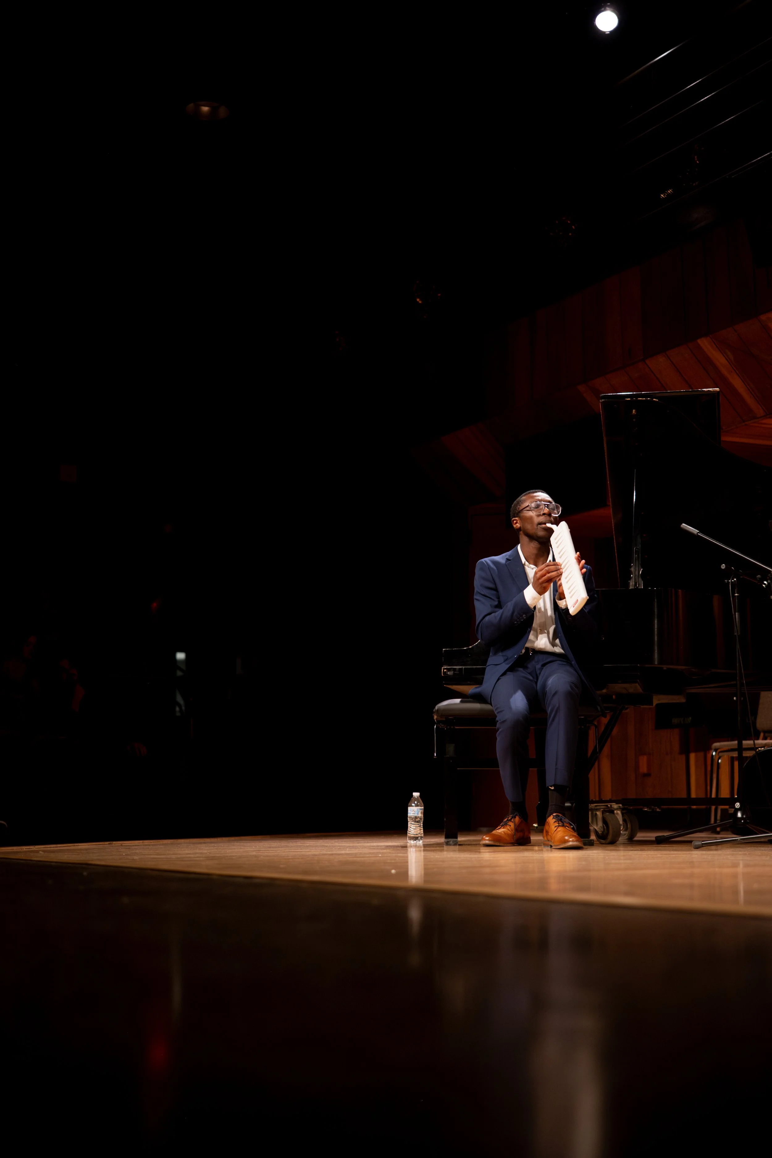 A man in a dark blue suit and brown shoes sits at a black grand piano on stage, holding a paper and speaking into a microphone. A water bottle is on the floor beside him, and a stage light is shining from above.