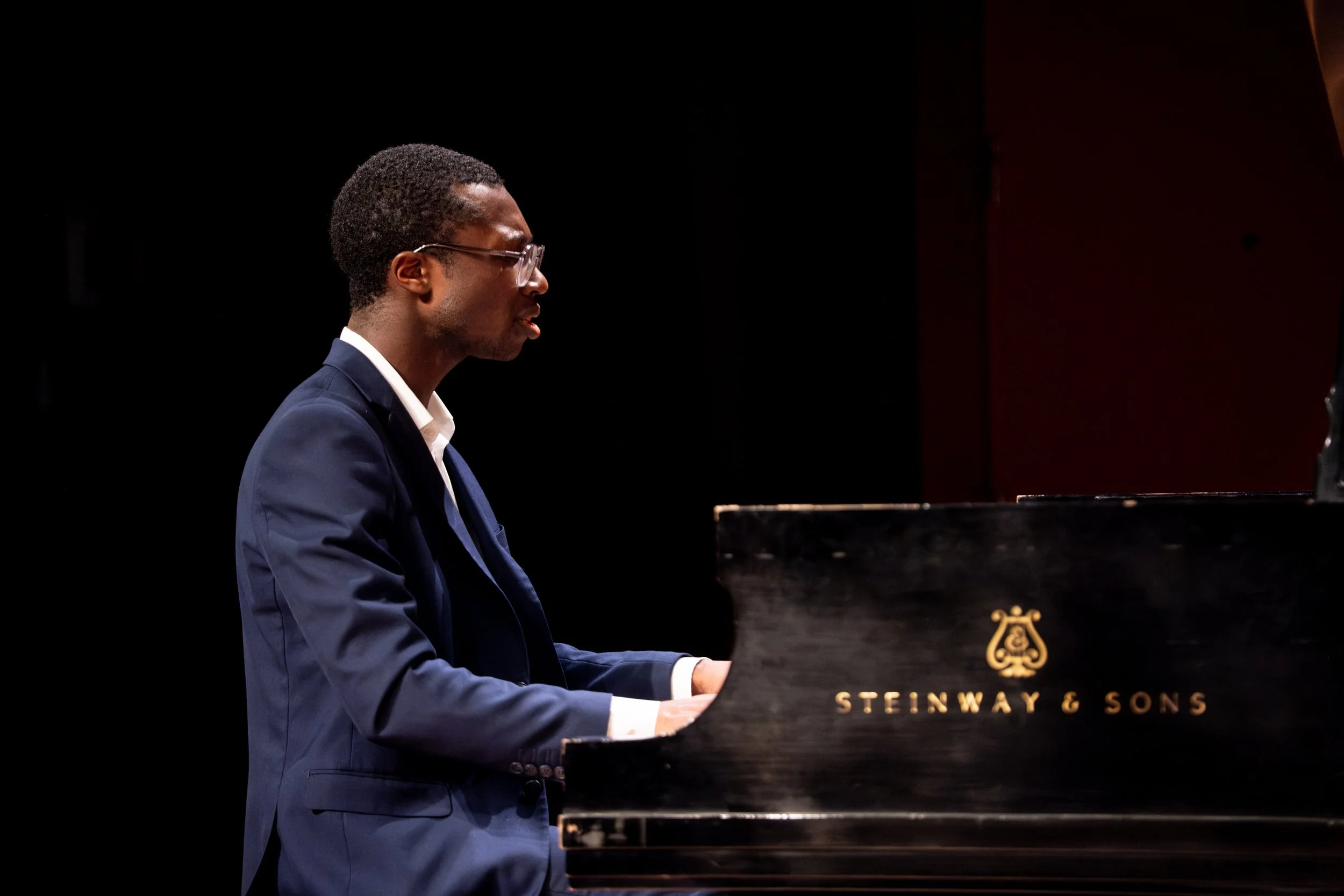 A man wearing glasses and a blue suit plays a grand piano on a dark stage.