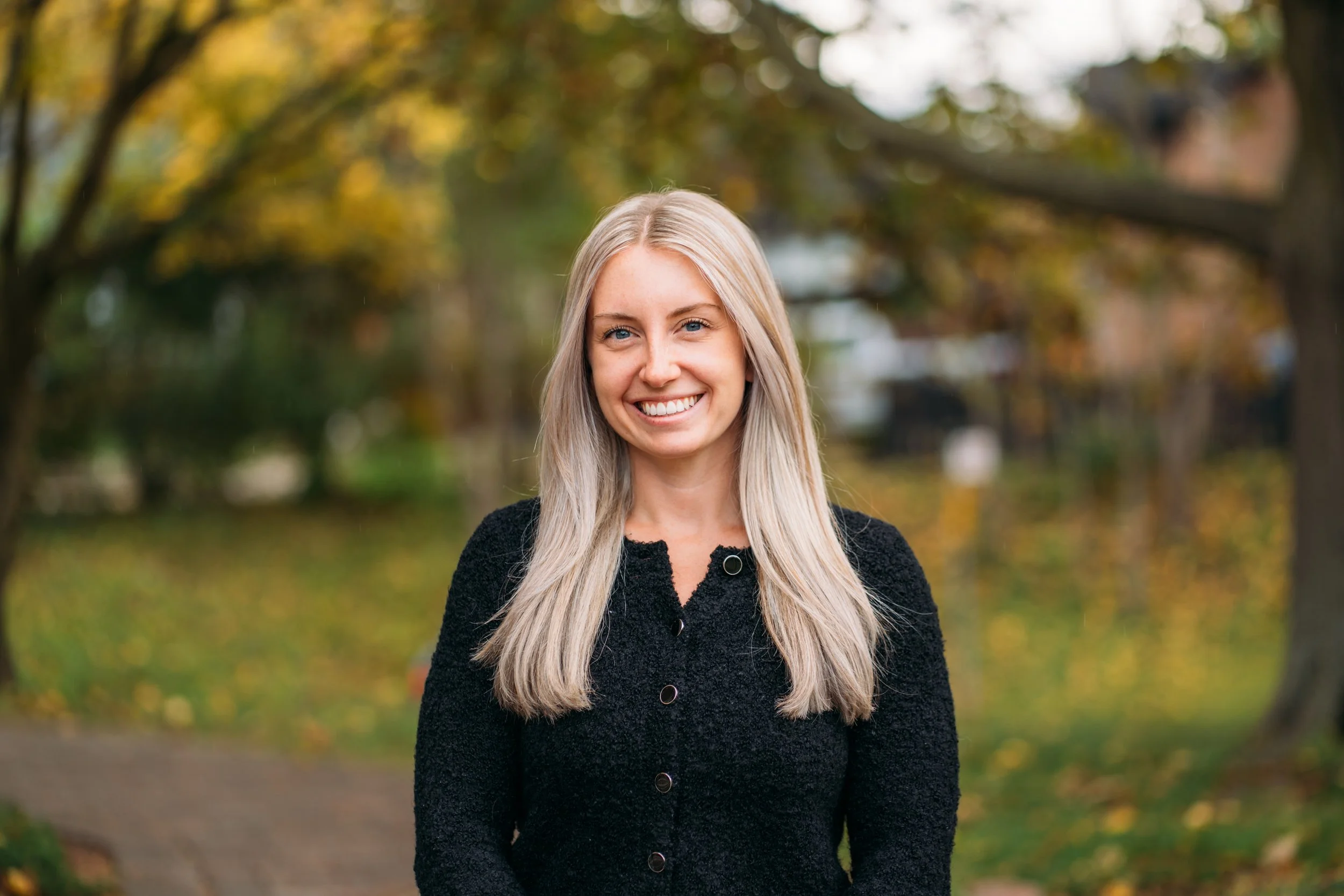 A smiling blonde woman in a black coat standing outdoors in a park with autumn trees in the background.
