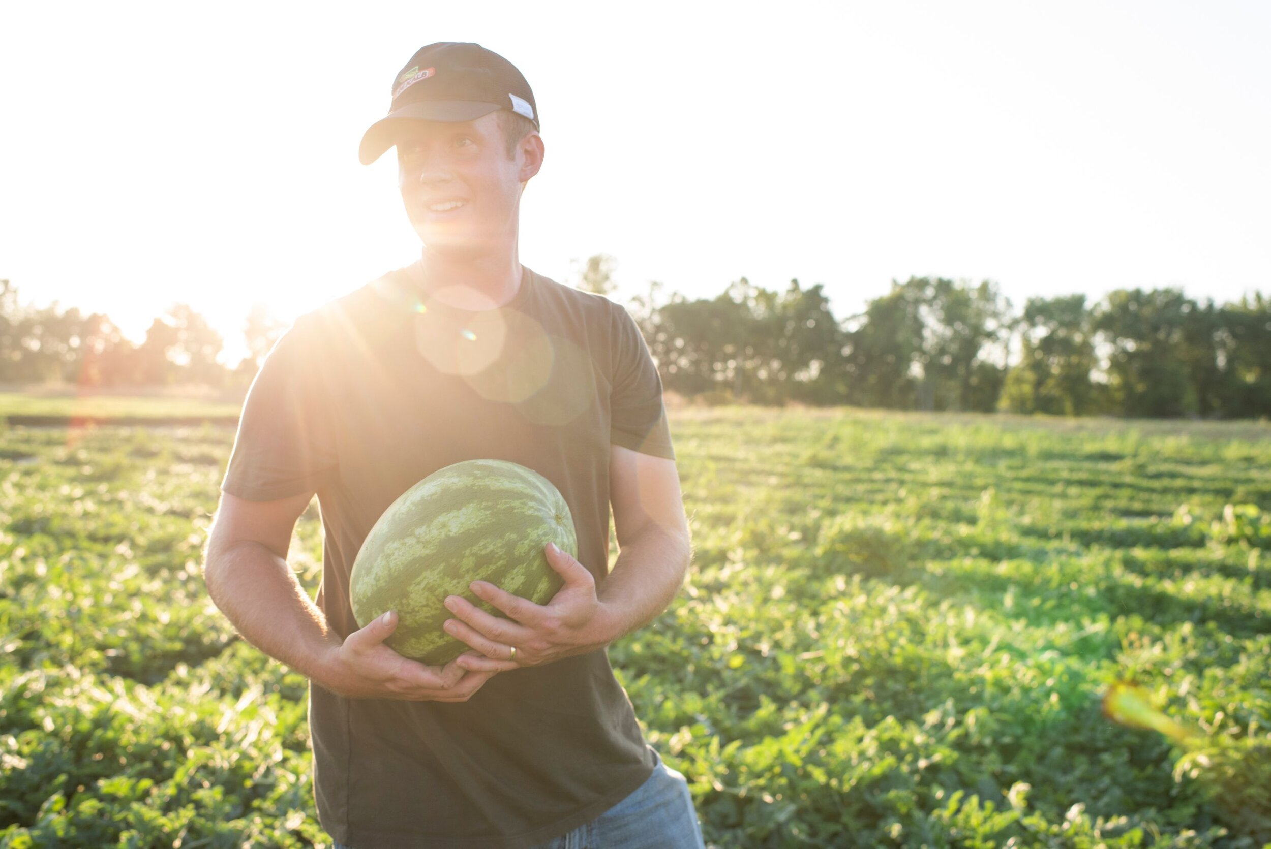 A high school boy in jeans and a cap holding a watermelon in a green field