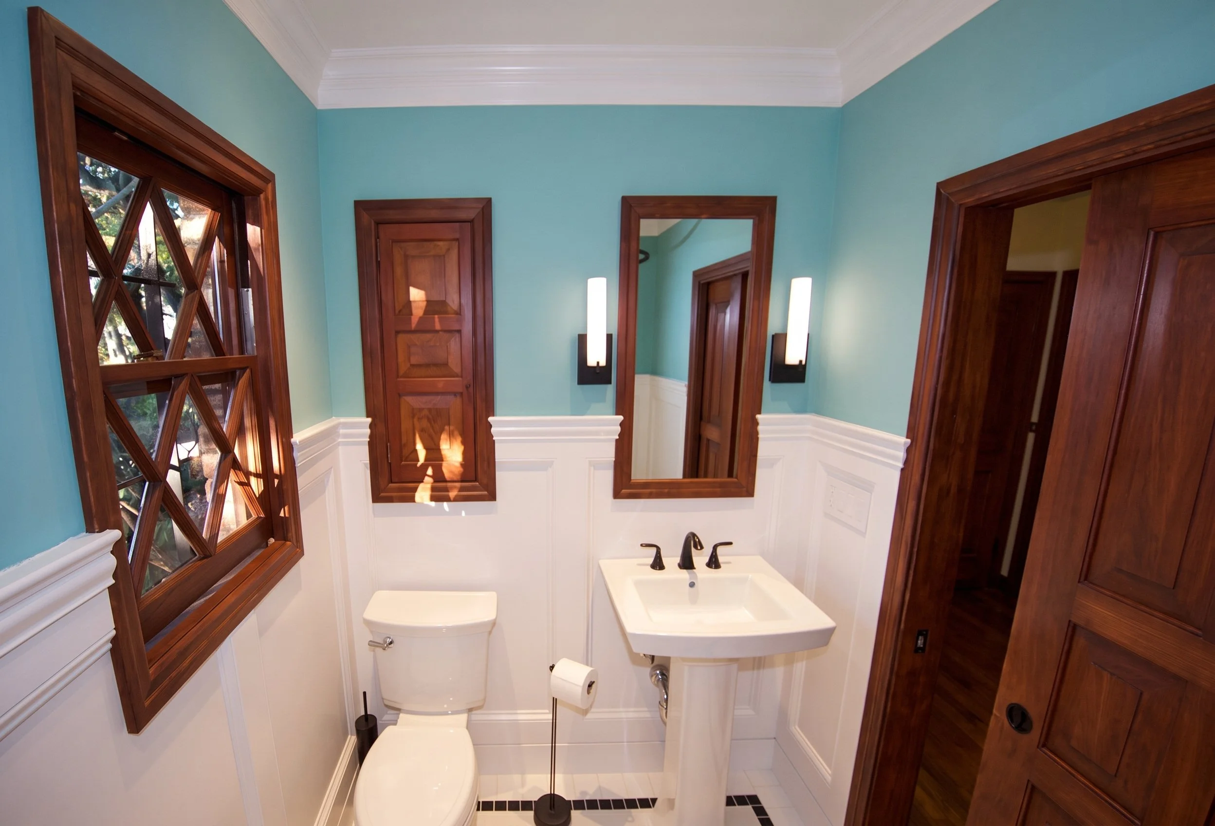 Pedestal sink and toilet below the wood-framed mirror and original window.