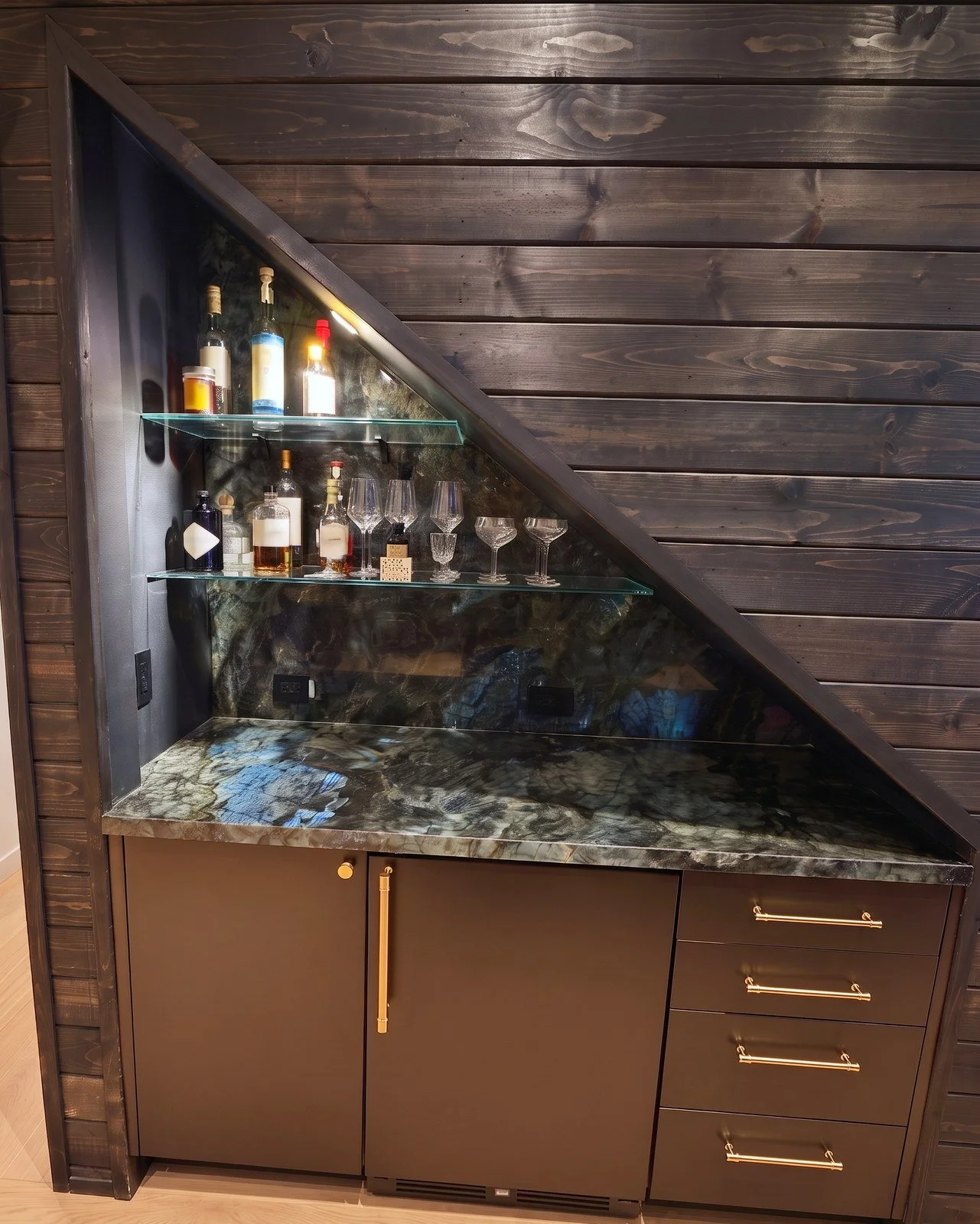 Built-in bar in a triangular niche under a stair, with glass shelves, a dark stone counter and brass cabinet pulls.