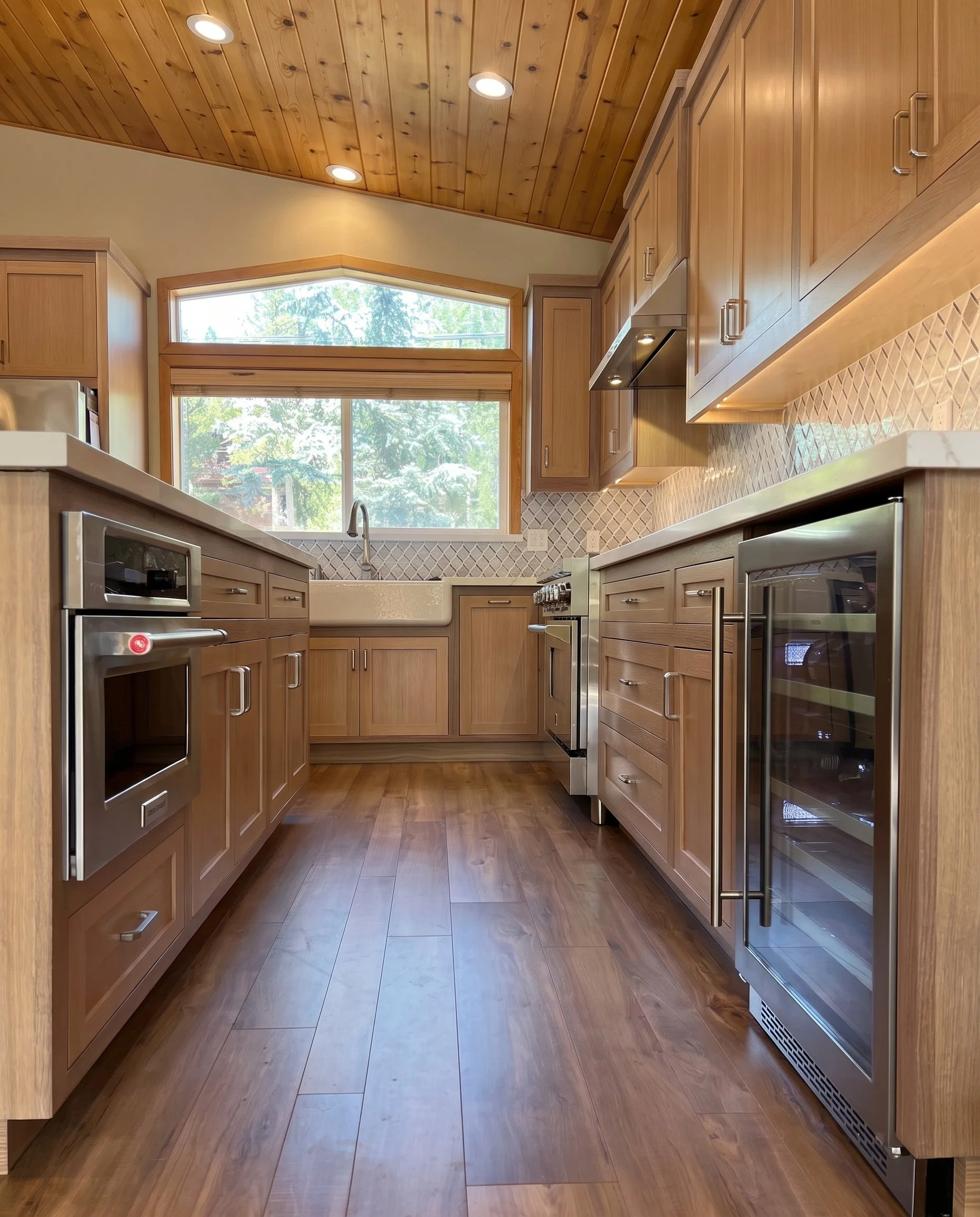 View down the kitchen toward the window, with wall ovens and a wood plank ceiling.
