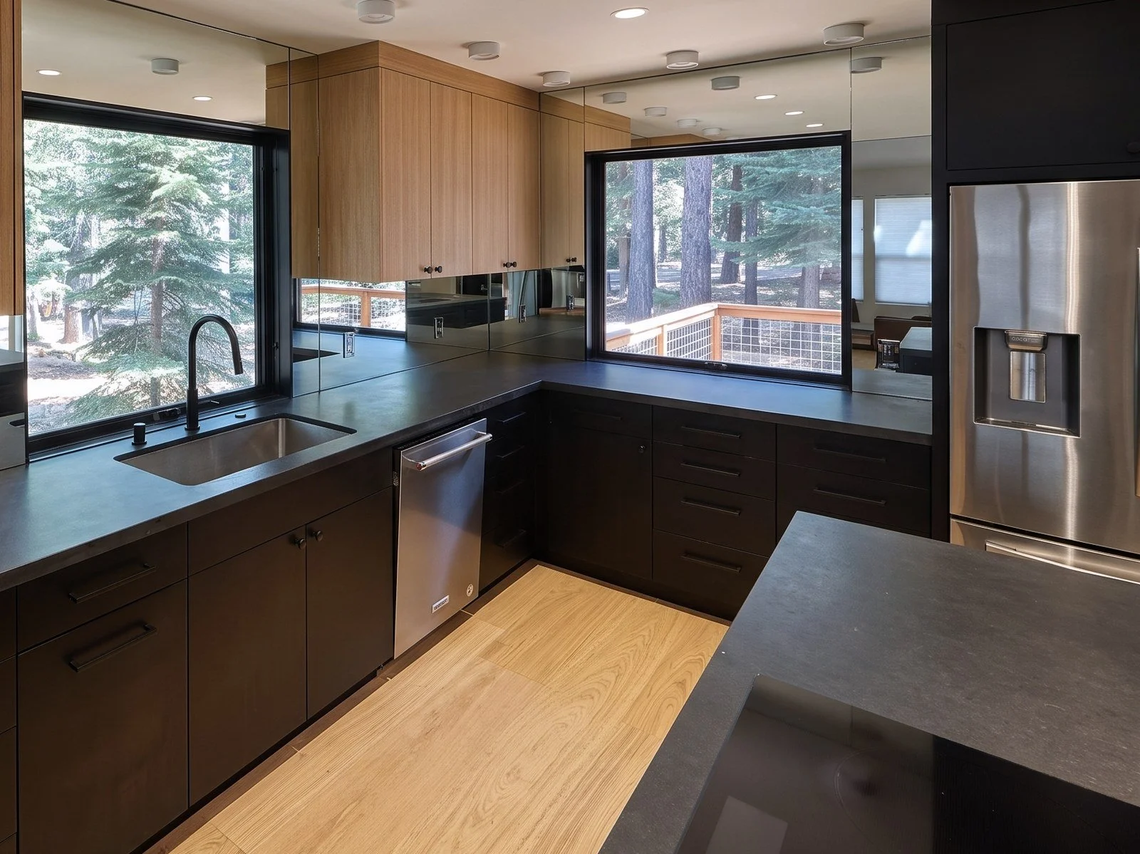 Kitchen counter run with oak upper cabinets, black lower cabinets, a mirror backsplash and two picture windows onto pine trees.