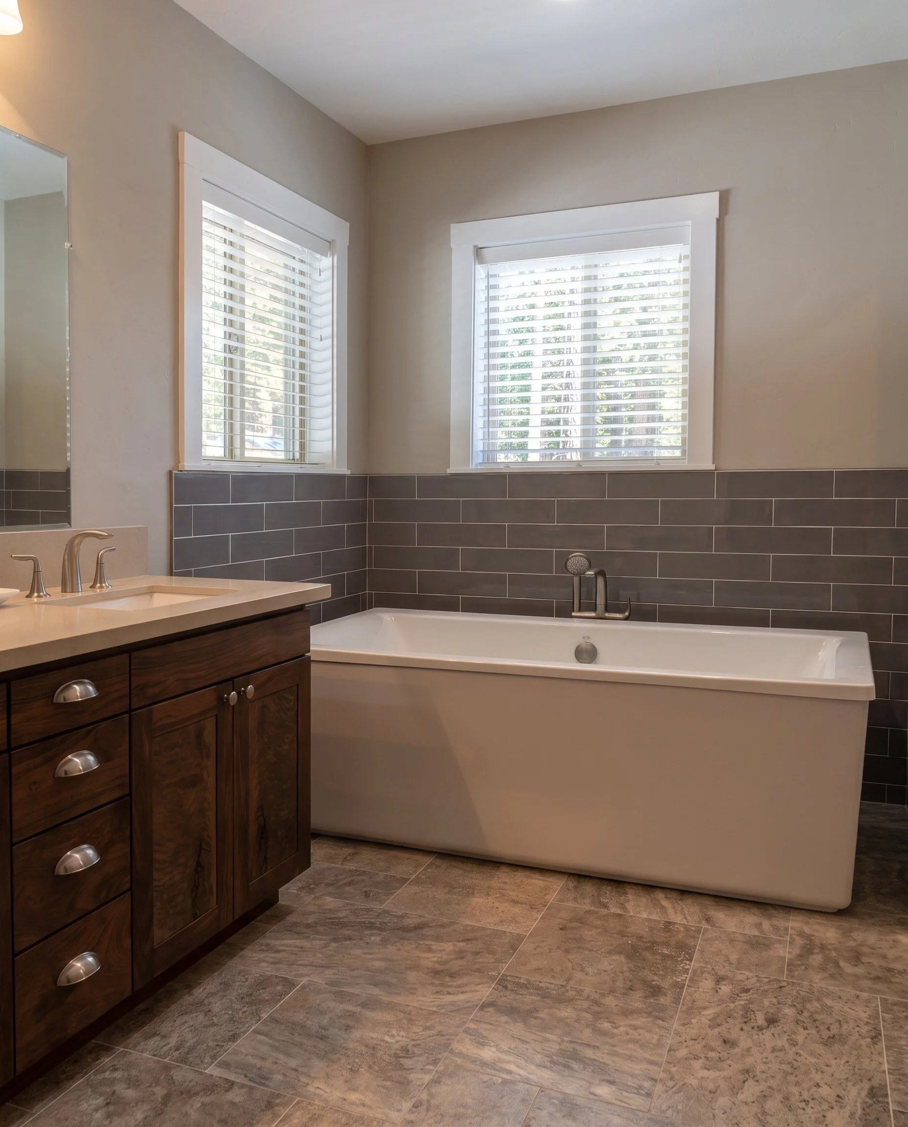 Freestanding soaking tub under shuttered windows, with gray subway tile wainscot and a dark wood vanity.