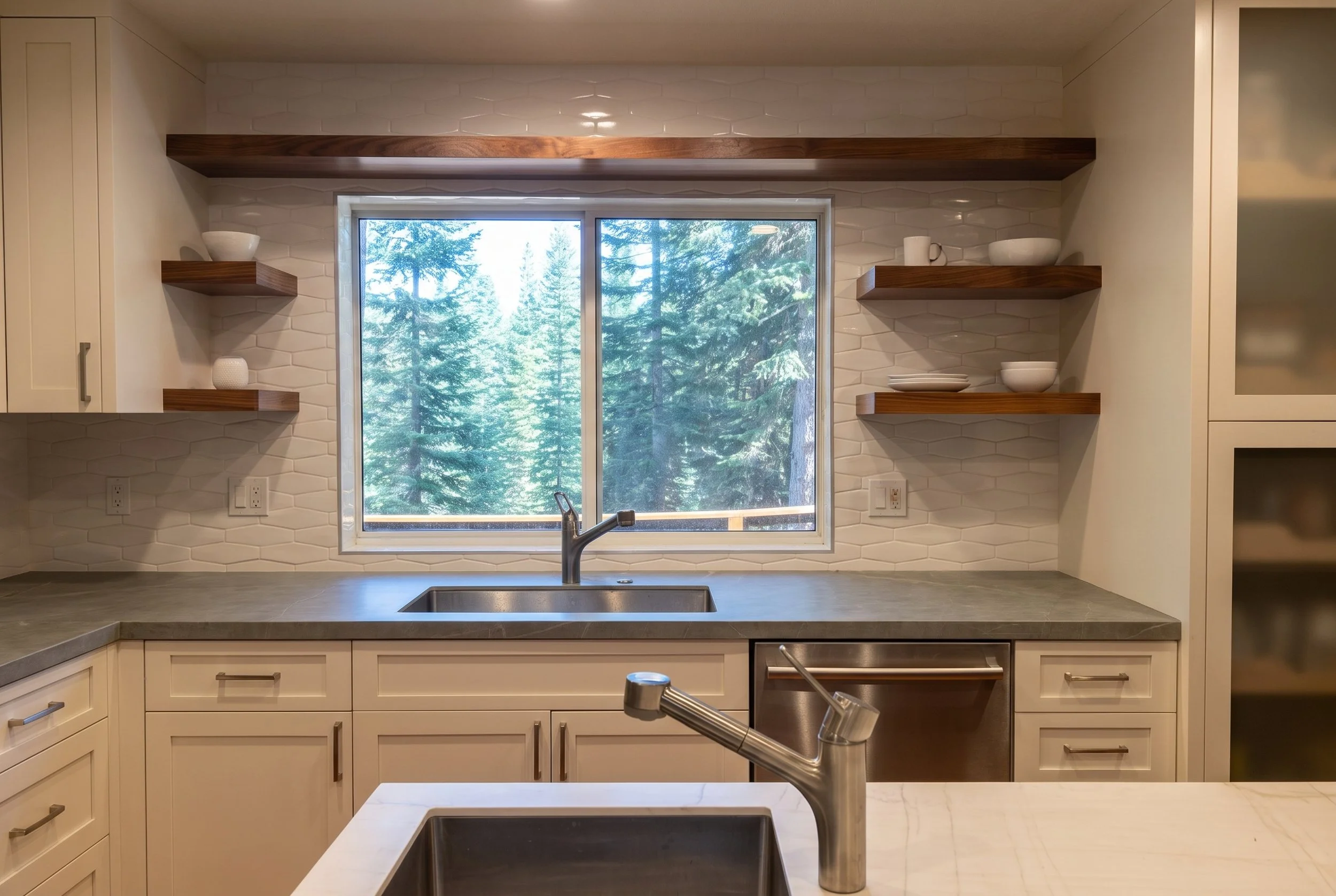 Sink wall with gray stone counters and wood floating shelves on both sides of the window.