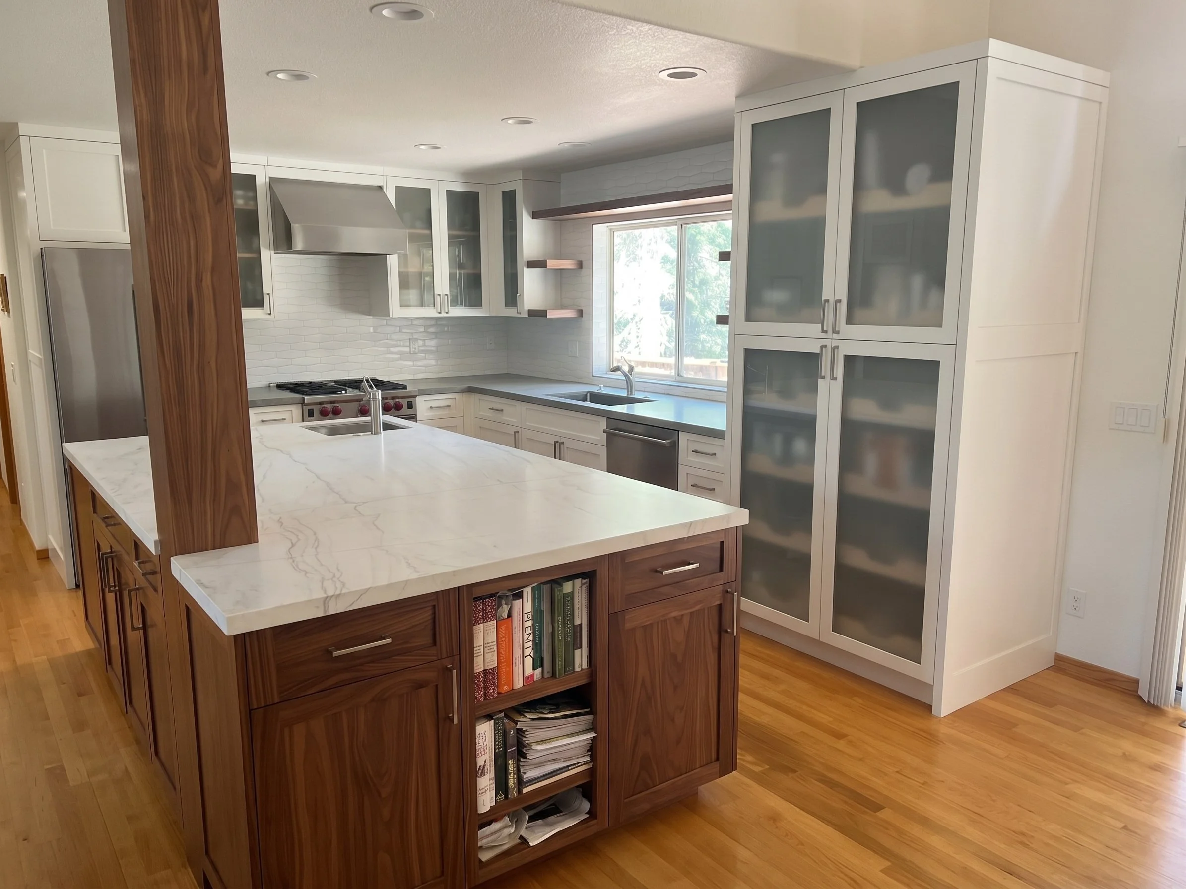 Dark wood island with a white stone top and an end bookshelf, beside tall frosted glass cabinets.