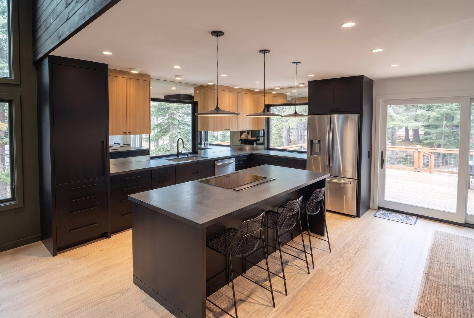 Kitchen with black cabinets, oak upper cabinets, a dark stone island with three stools and three pendant lights.