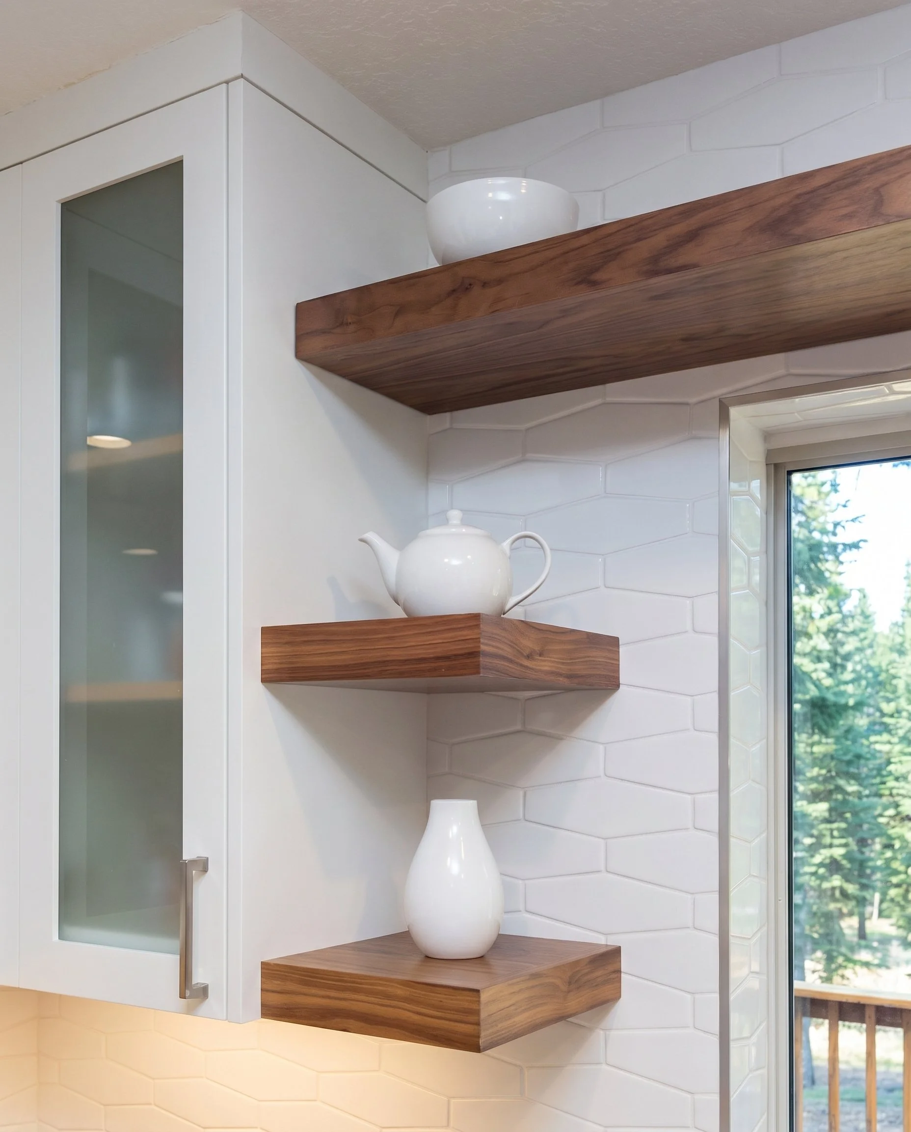Detail of the wood floating shelves against white tile, beside a glass-front cabinet.