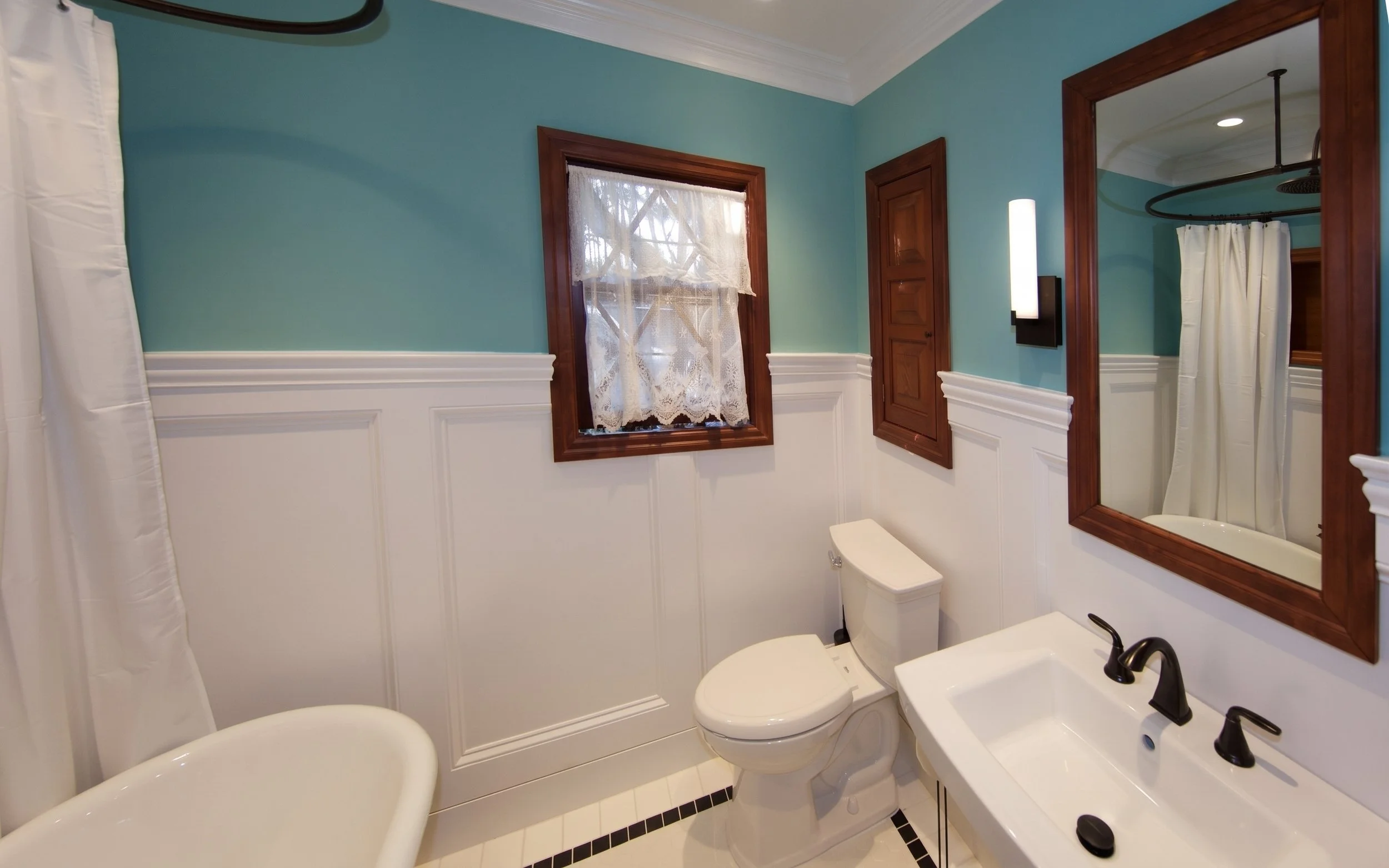 Clawfoot tub, pedestal sink and wood-framed mirror above white paneled wainscot.