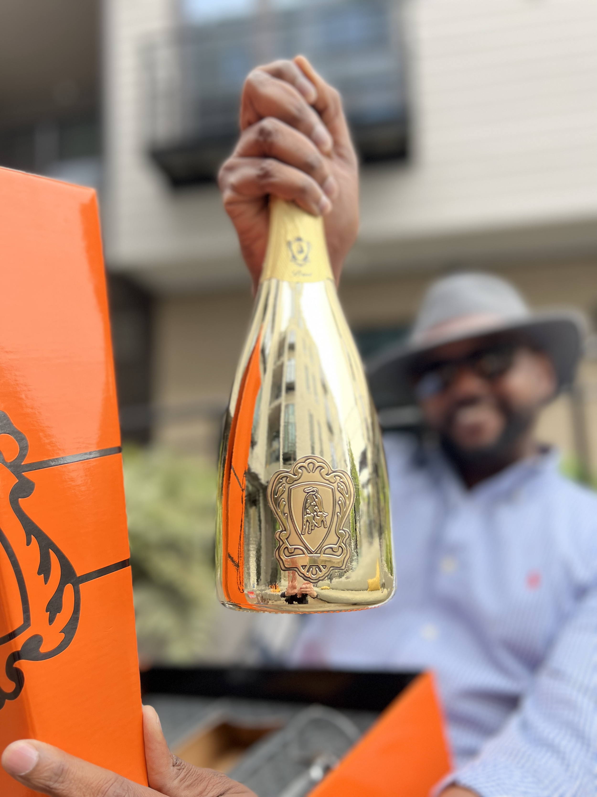 A person holding a gold-colored bottle of Veuve Clicquot champagne with an orange box nearby, and a man wearing a hat and glasses smiling in the background.