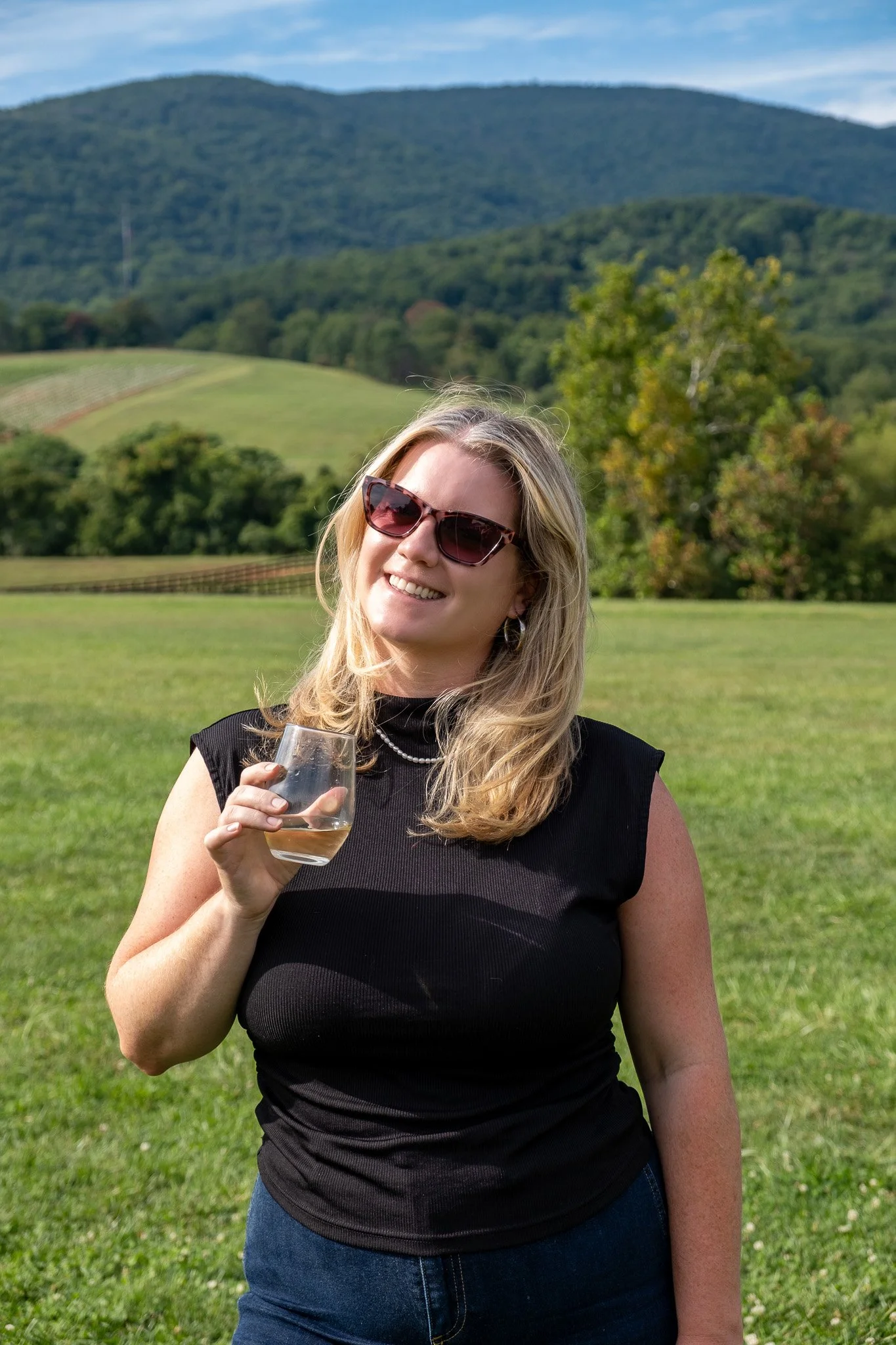 A woman with blonde hair wearing sunglasses, a black sleeveless top, and a pearl necklace, holding a glass of white wine outdoors on a grassy field with rolling hills and trees in the background.