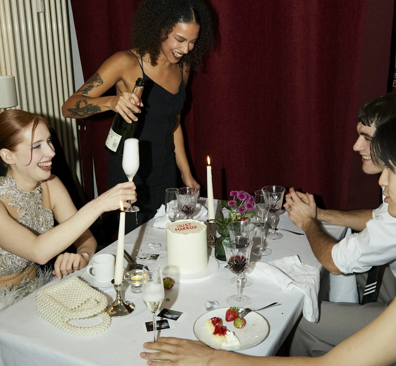 A woman is pouring champagne for a woman celebrating her marriage with a cake that says 'Just Married,' at a table with friends, strawberries, flowers, and candles.