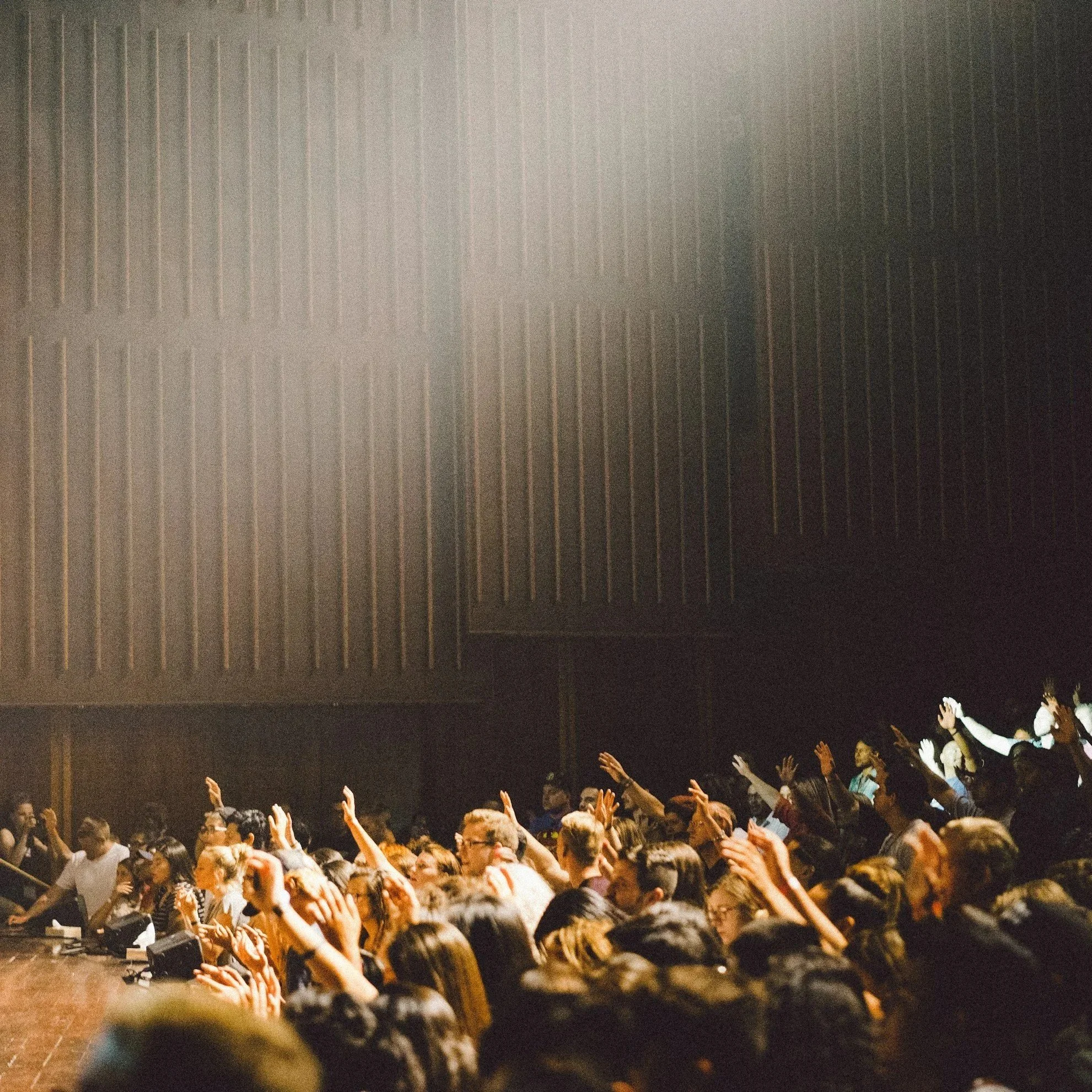 Crowd of people worshipping with many raising their hands, illuminated by stage lights.