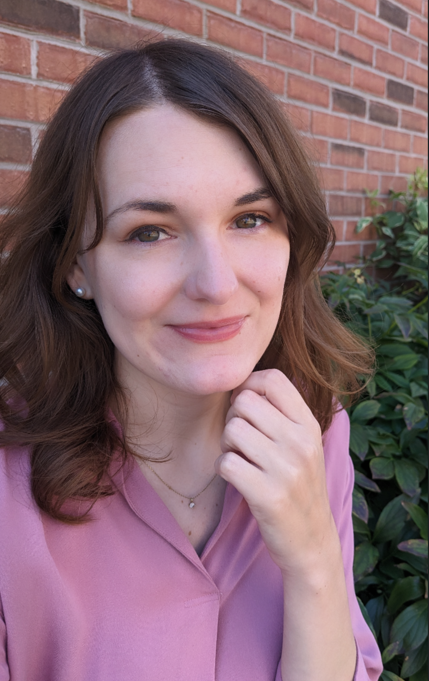 A therapist with shoulder-length brown hair, wearing a pink blouse, smiling and resting her hand on her chin, standing in front of a brick wall and green foliage.