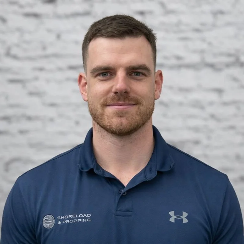 A man with short brown hair and a beard, wearing a navy blue Under Armour polo shirt with 'SHORELOAD & PROPPING' embroidered on it, standing in front of a light gray brick wall.
