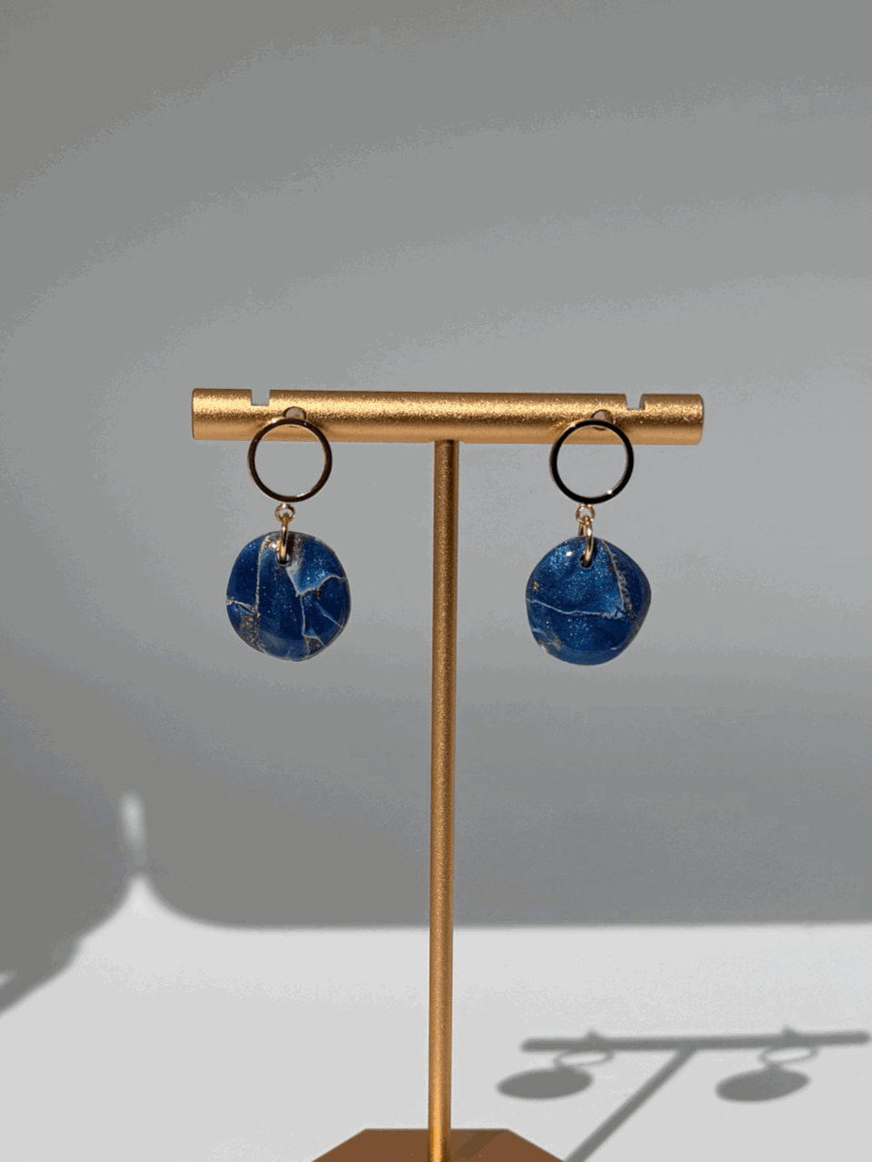 Pair of earrings with blue marble-like pendants hanging from black circular hooks displayed on a gold stand.