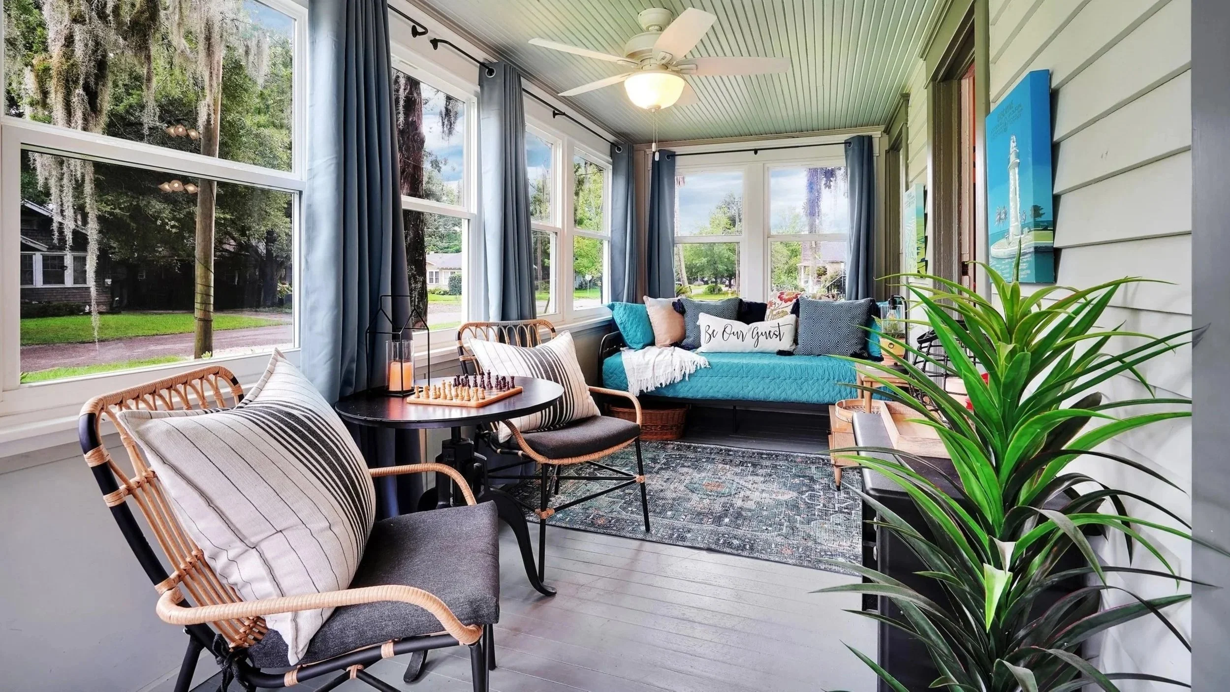 Bright, enclosed sunroom featuring blue curtains, a cozy daybed with teal bedding, two woven accent chairs around a small table set with a chessboard, a white ceiling fan, and large windows overlooking trees draped with Spanish moss.