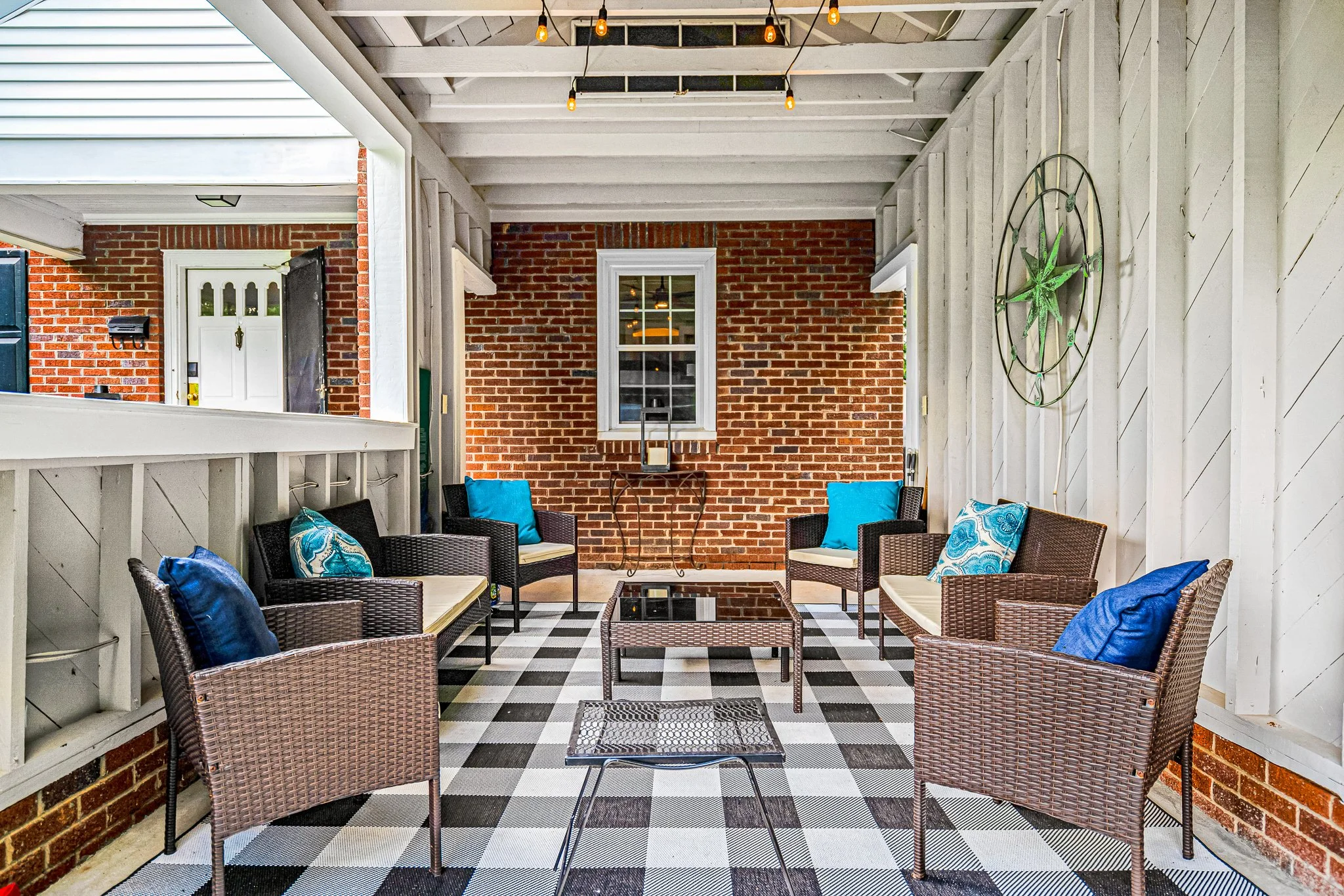Covered brick patio featuring a brown wicker outdoor conversation set with bright blue accent pillows, a black and white plaid rug, and a metal compass wall hanging.