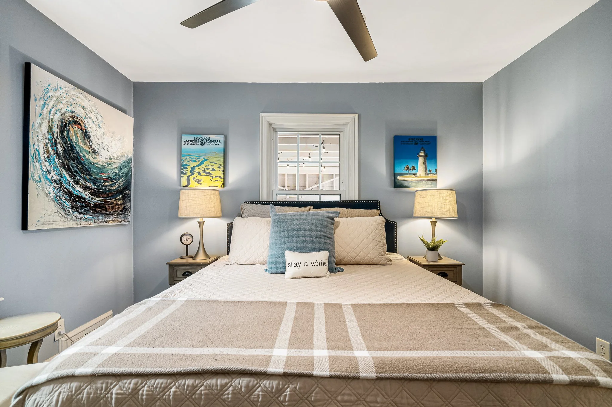 Cozy coastal bedroom interior featuring a bed with blue and beige pillows, a 'stay a while' accent pillow, matching bedside lamps, and ocean-themed wall art.