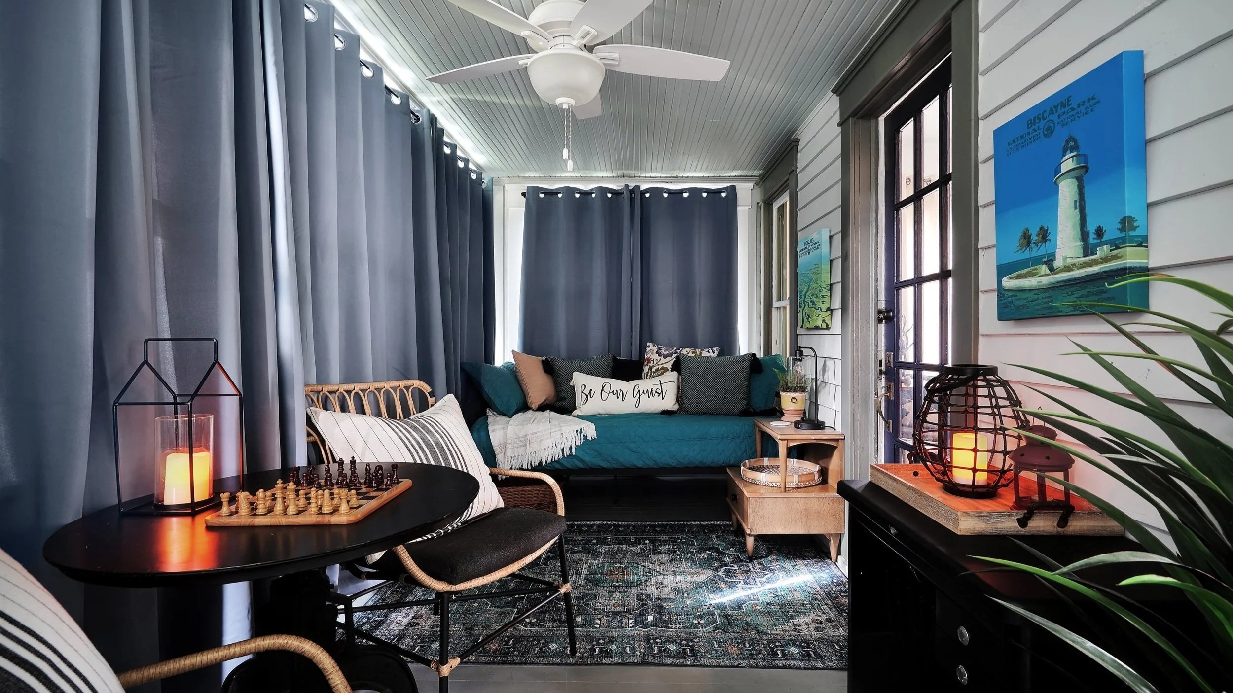 Enclosed sunroom lounge featuring full-length blue privacy curtains, a round chess table with a glowing lantern candle, a teal daybed with decorative pillows, and a blue lighthouse wall art print.