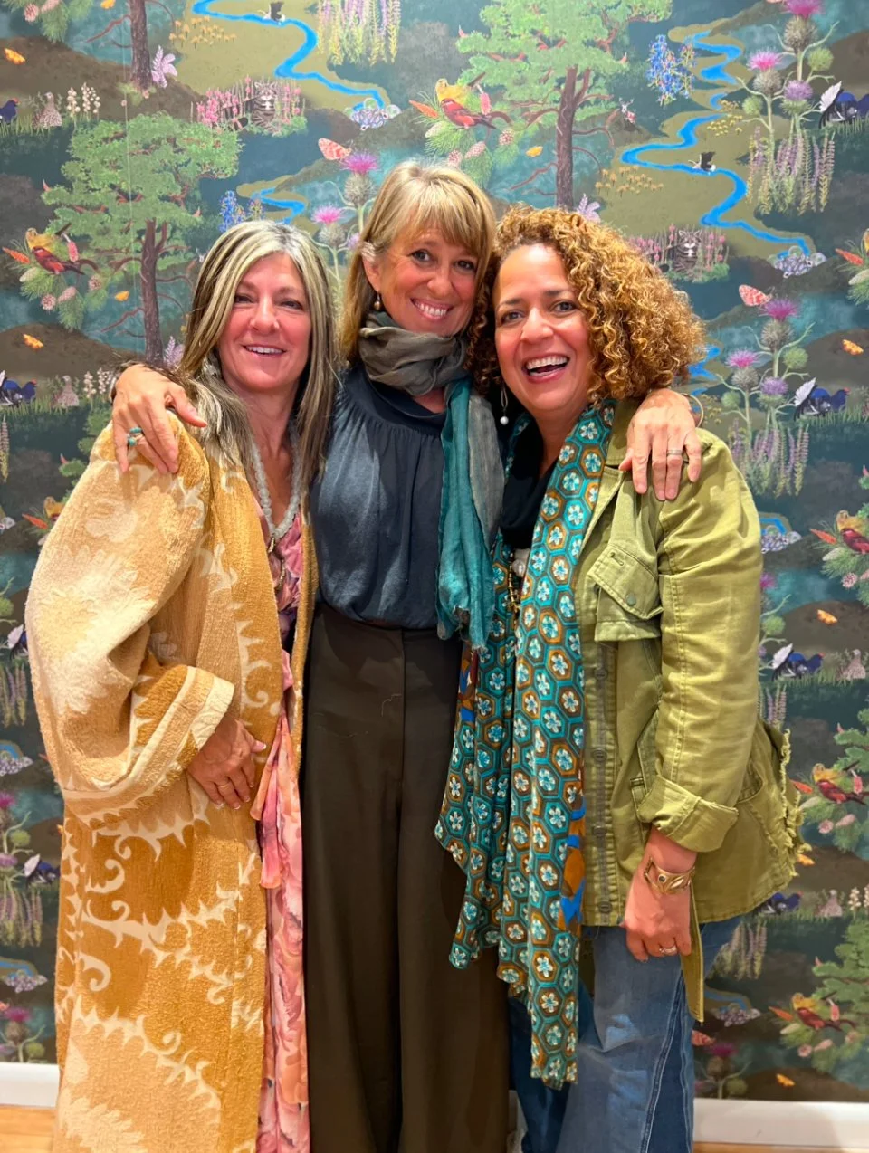 Three women happily posing in front of a vibrant, nature-inspired wallpaper, dressed in stylish, textured clothing that complements the sophisticated and cozy interior design setting.