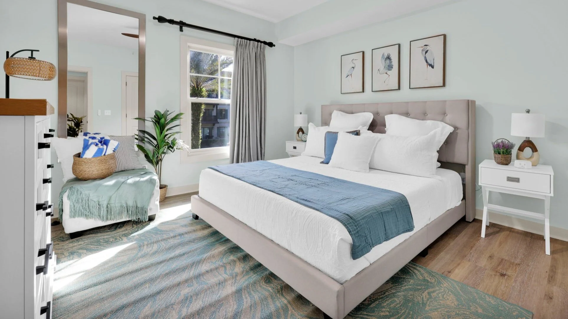 A bright and airy bedroom featuring a plush tufted king-sized bed with crisp white linens, accented by a soft blue throw and matching decorative pillows. The room is styled with a coastal-inspired palette, including a wave-patterned area rug.