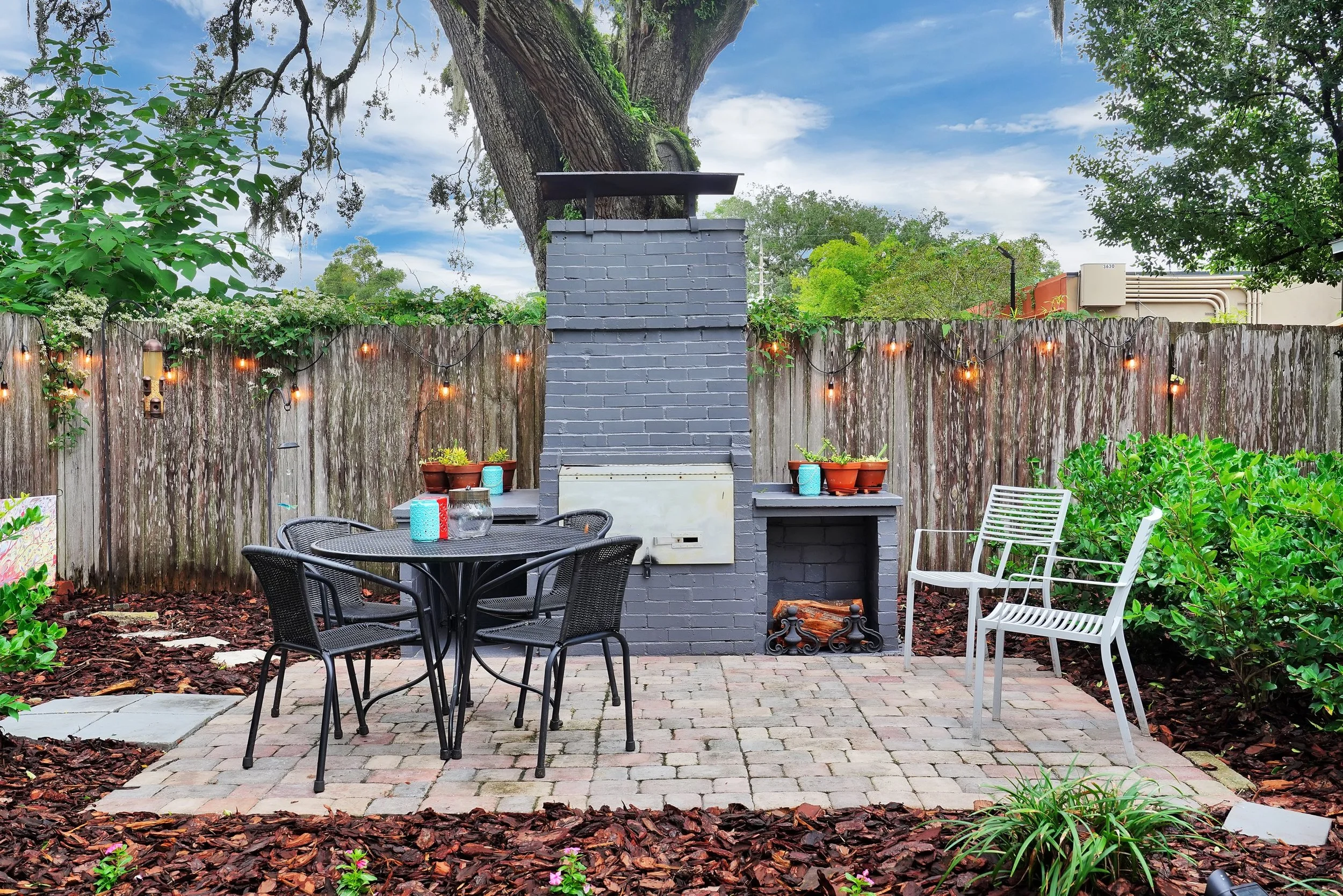 Backyard paver patio featuring a tall grey painted brick fireplace, a black metal dining set, white slatted chairs, and warm string lights hanging along a wooden privacy fence.