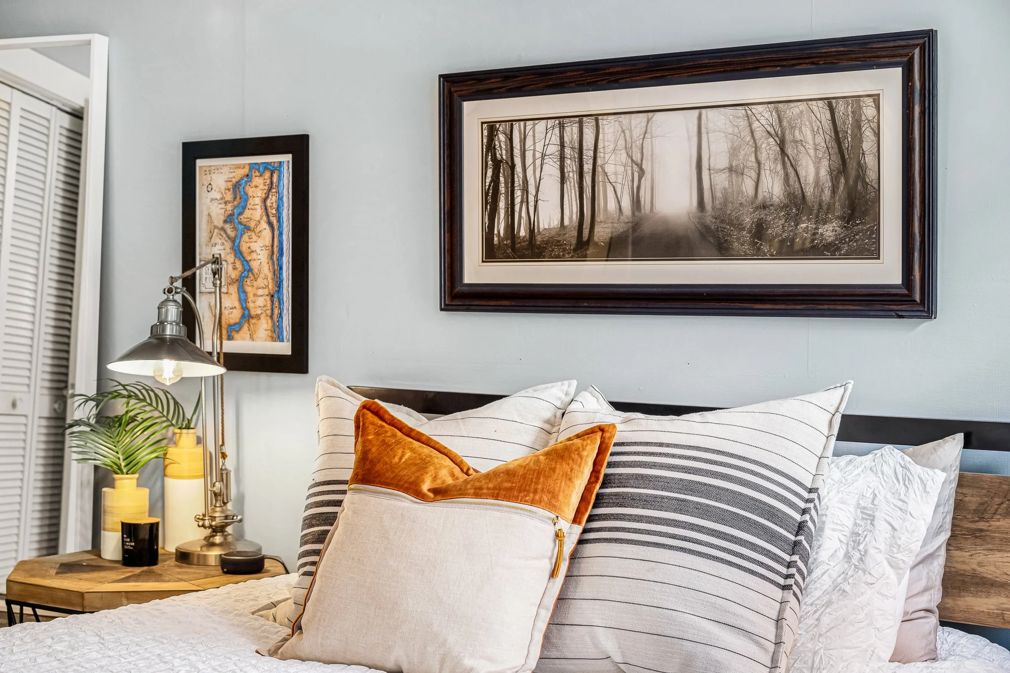 Cozy bedroom styling featuring a bed with striped and burnt orange velvet pillows, an industrial metal reading lamp, and a large framed misty forest photograph.