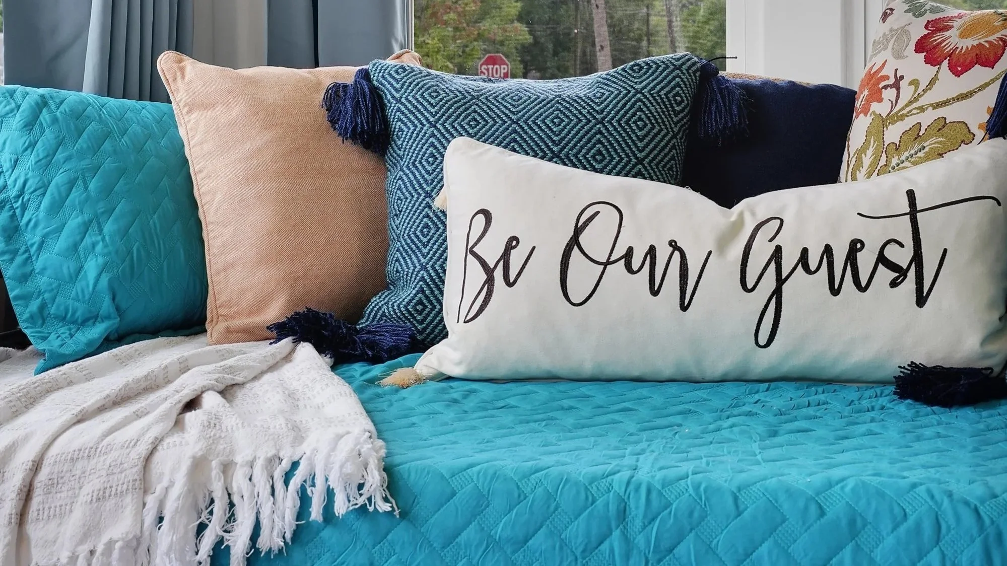 Close-up of a cozy daybed arranged with colorful throw pillows, featuring a white lumbar pillow in front with black cursive text reading "Be Our Guest," set against teal, peach, blue geometric, and floral patterned cushions.