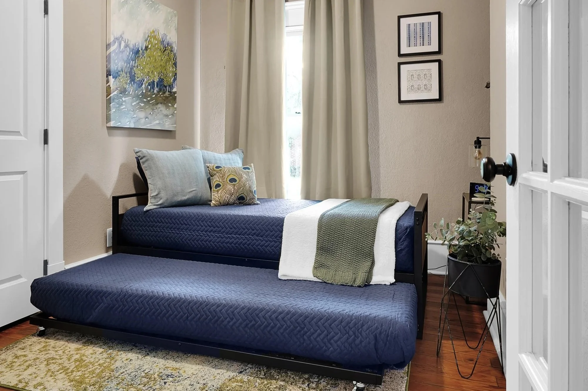 Wide view of a cozy guest bedroom seen through a doorway, showing a black metal daybed with its lower trundle bed pulled out, both featuring matching navy blue quilted bedding, light blue pillows, and framed wall art.