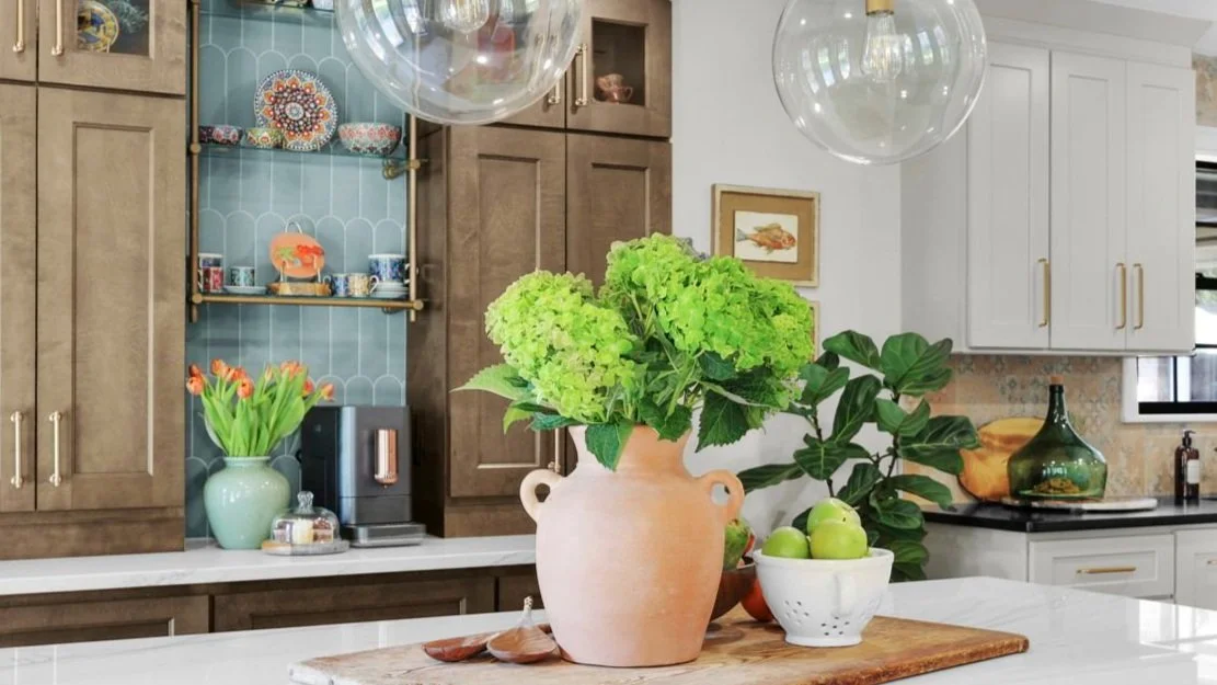 Kitchen coffee bar with blue scalloped tile backsplash, warm wood cabinets, and terracotta vase of green hydrangeas on a marble island.