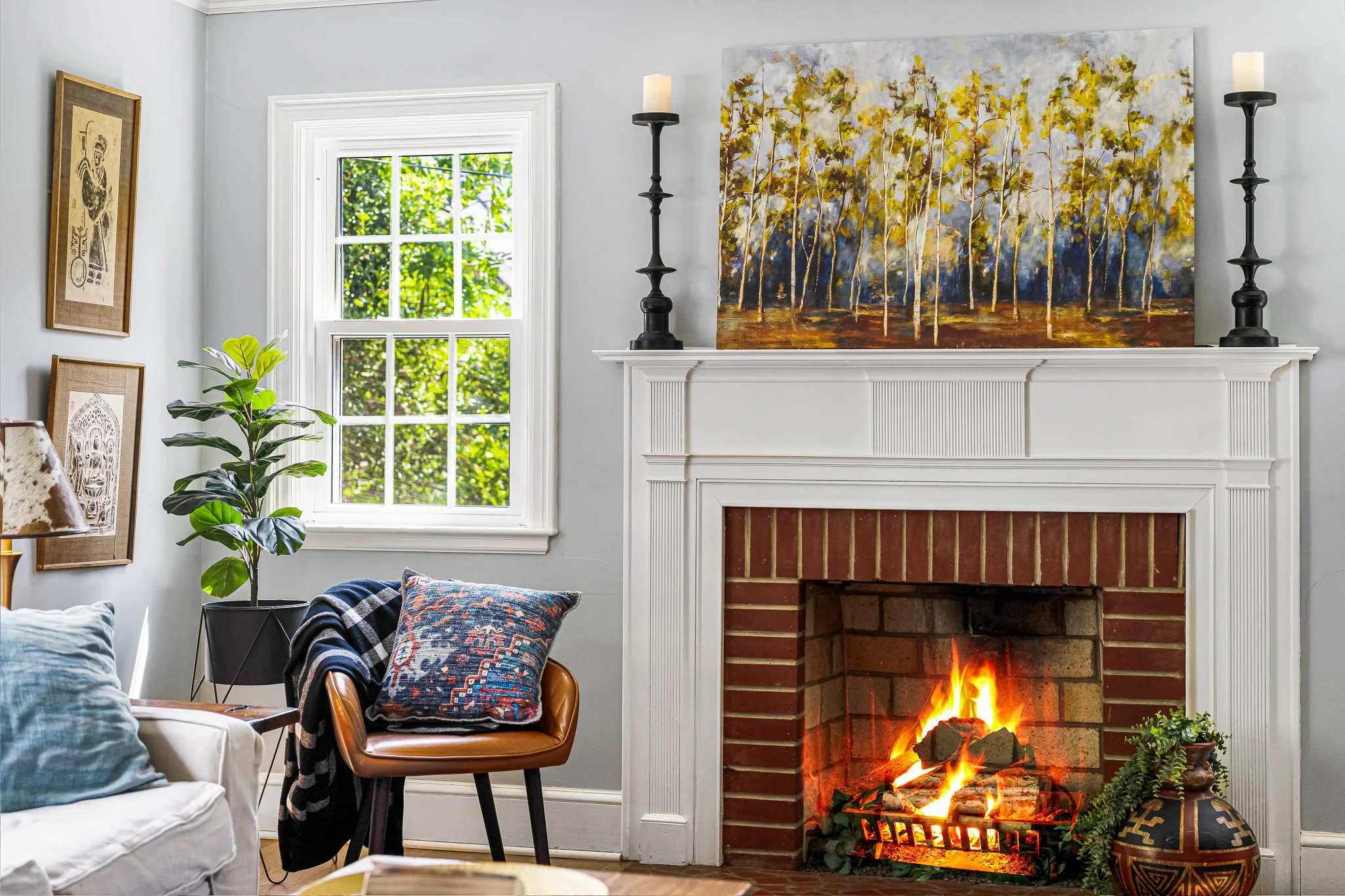 Elegant white fireplace mantel decorated with a large landscape tree painting, tall black candlesticks, a window, and a potted fiddle leaf fig plant.