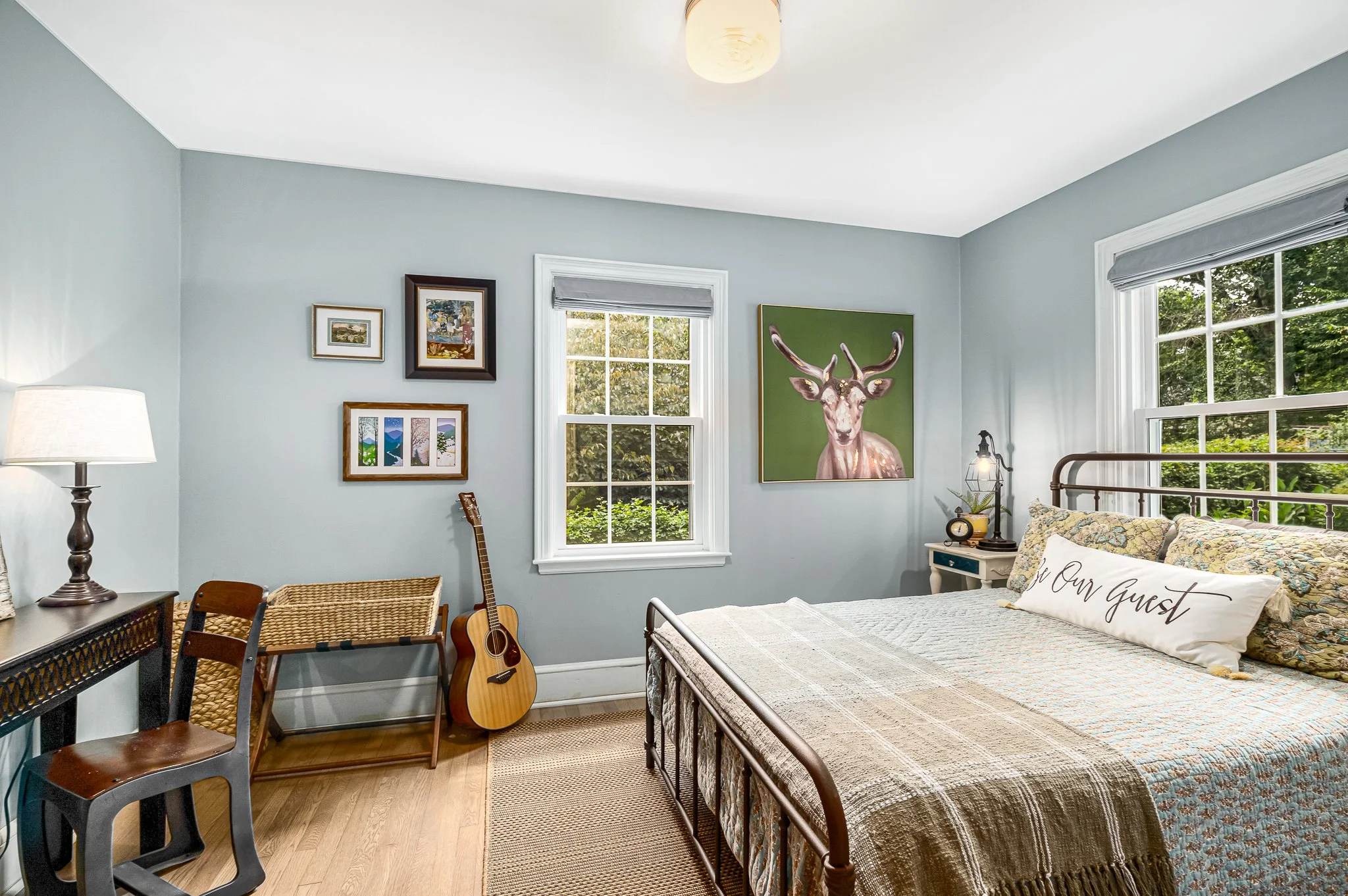 Cozy guest bedroom interior with a metal bed frame, a 'Be Our Guest' accent pillow, a large rustic deer painting, and an acoustic guitar leaning against light blue walls.