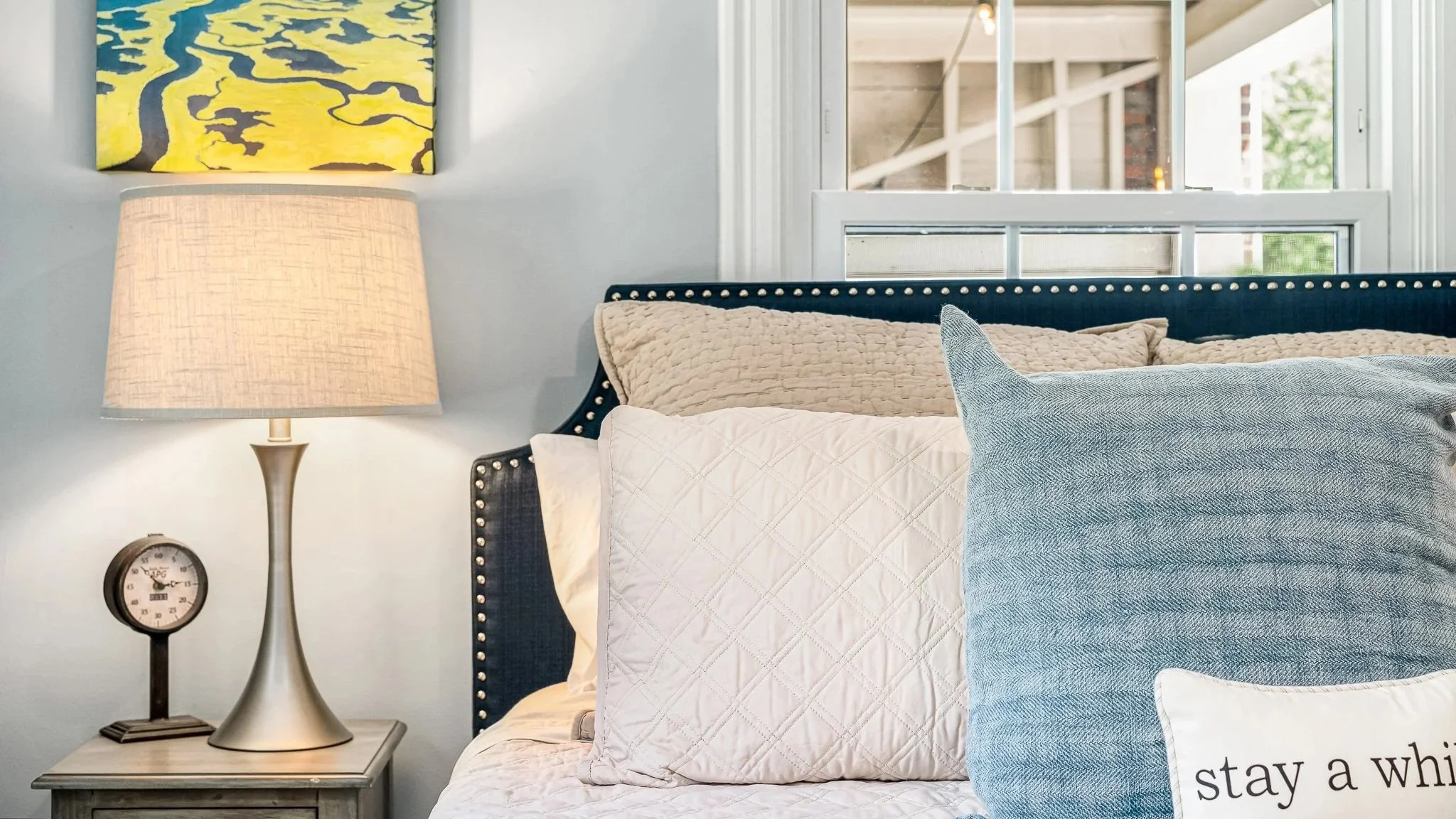 Close-up bedroom details showing a blue studded headboard, textured blue and beige pillows, and a modern silver lamp next to a vintage clock on a nightstand.