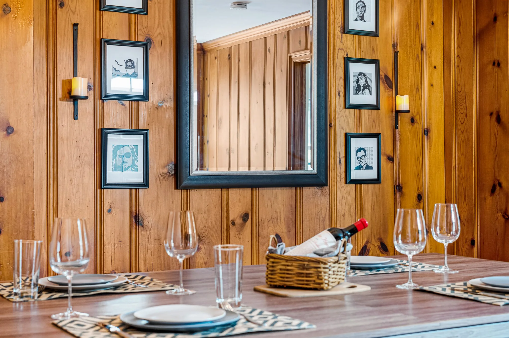 Rustic dining room table setting with wine glasses, plates, and a bottle of wine against a vertical wood-plank wall with a large mirror.