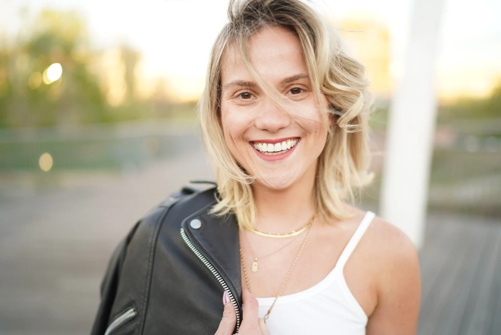 A smiling woman with blonde, wavy hair, wearing a white tank top, layered gold necklaces, and a black leather jacket draped over her shoulder, outdoors during daytime.