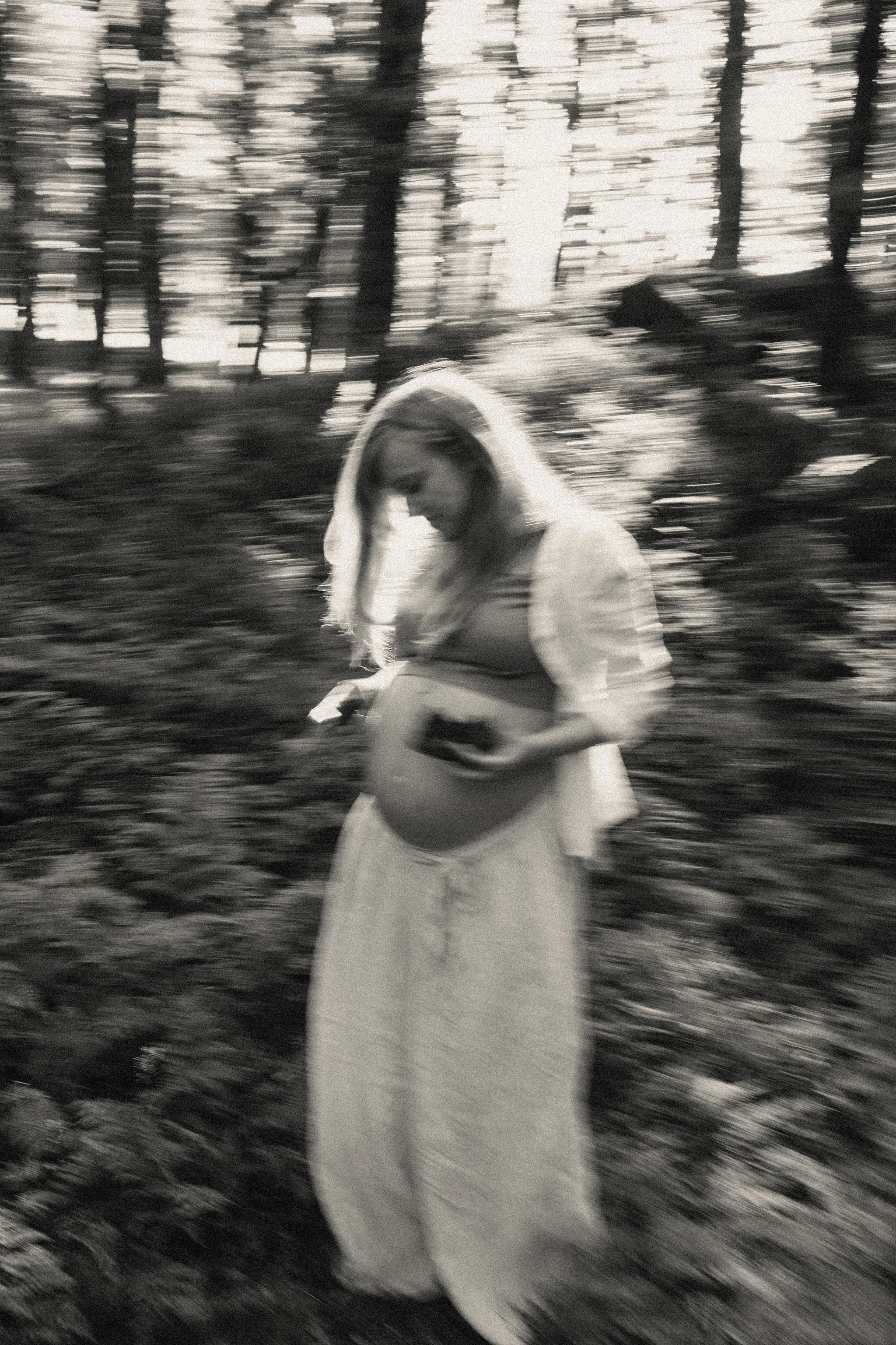 A woman with long hair, wearing a long skirt and a light-colored shirt, looks down at her pregnant belly while holding a phone in her hand, standing outdoors surrounded by trees and foliage.