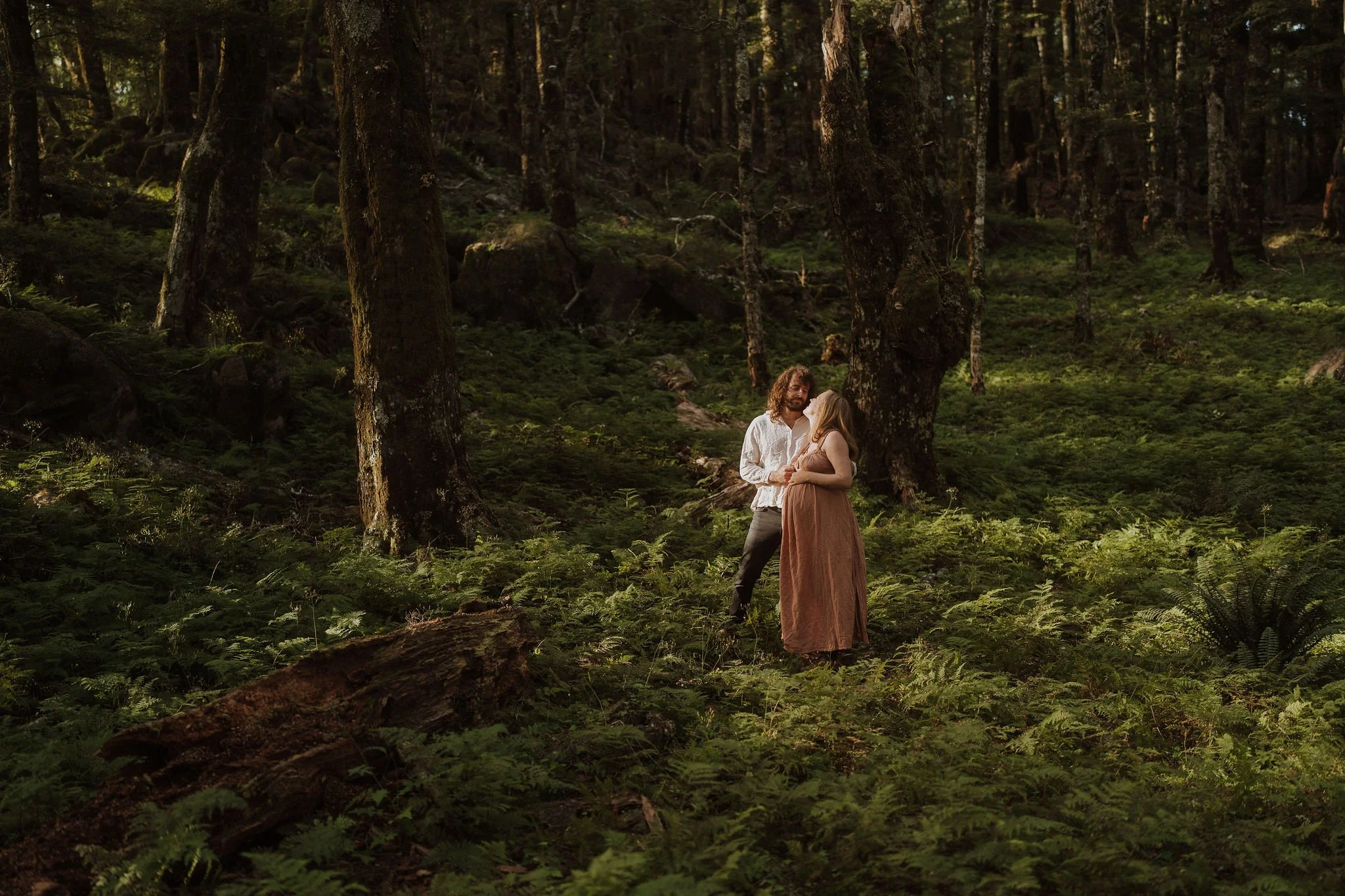 A couple standing in a lush, green forest during daytime, gazing into each other's eyes.