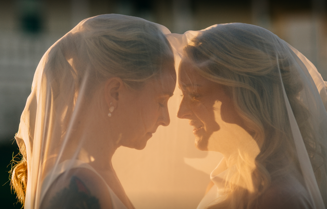 Two brides facing each other with veils, soft sunlight, intimate moment.