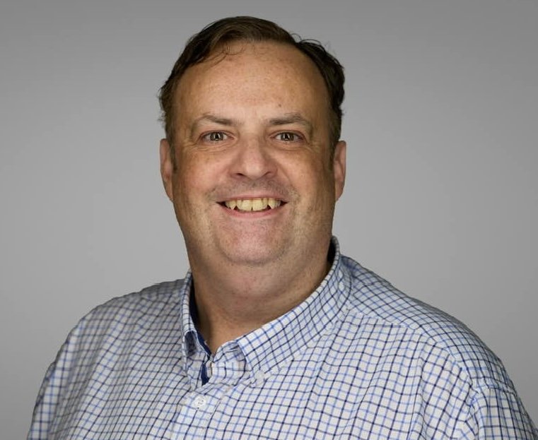 Ian smiling warmly at the camera. He has short brown hair and is wearing a light blue and white checkered button-up shirt. With a plain light grey background.