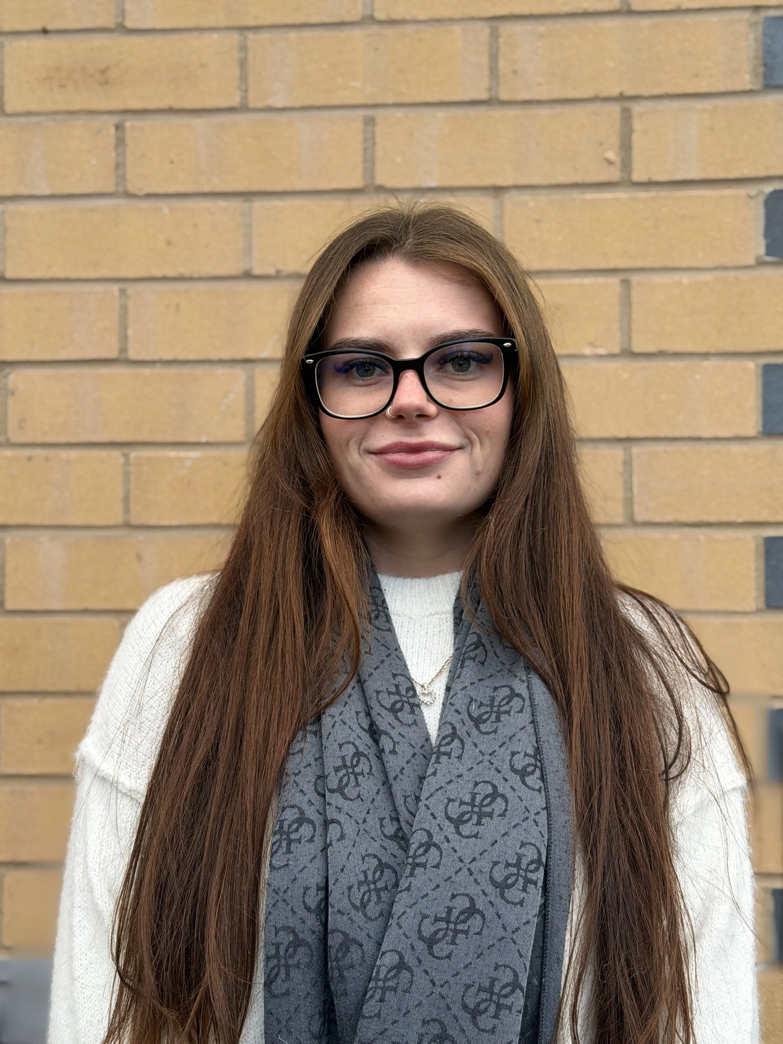 A young woman with long red hair, wearing glasses, a white sweater, and a gray patterned scarf, standing in front of a yellow brick wall.