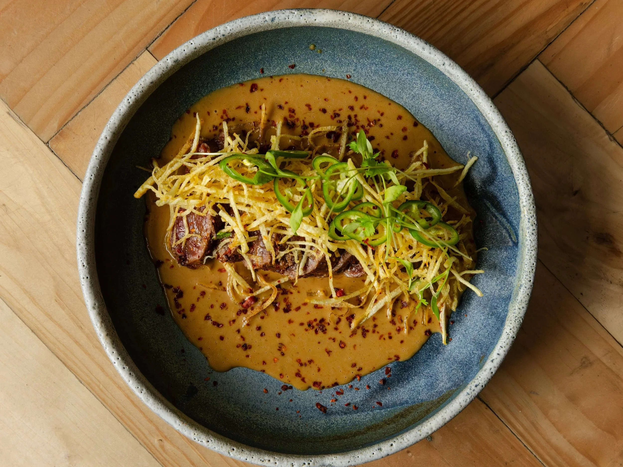 Venison tataki with shredded fried potatoes, green onions, beef, mustard sauce, and red pepper flakes on a wooden table.