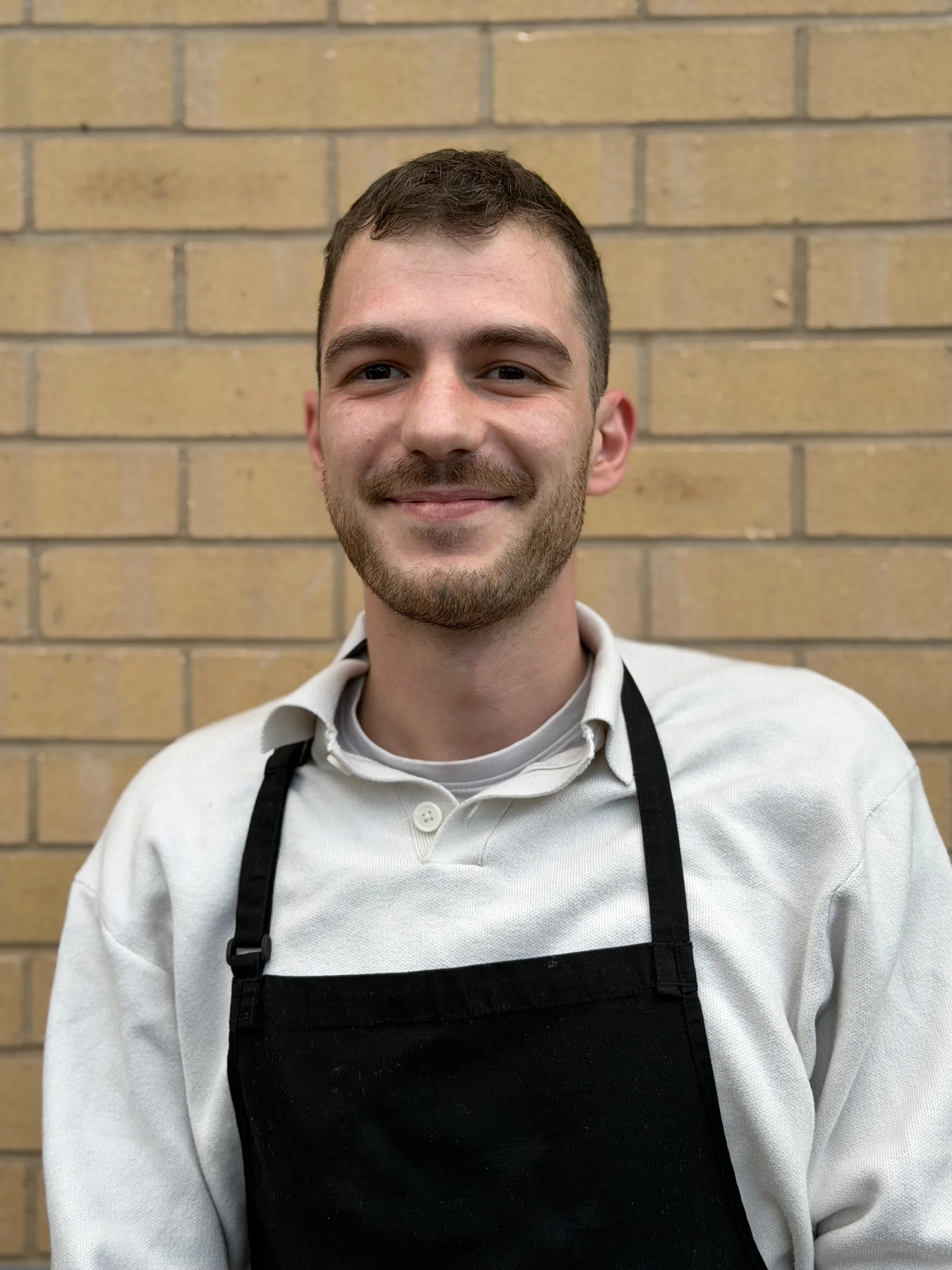 A smiling young man in a white shirt and black apron standing in front of a brick wall.