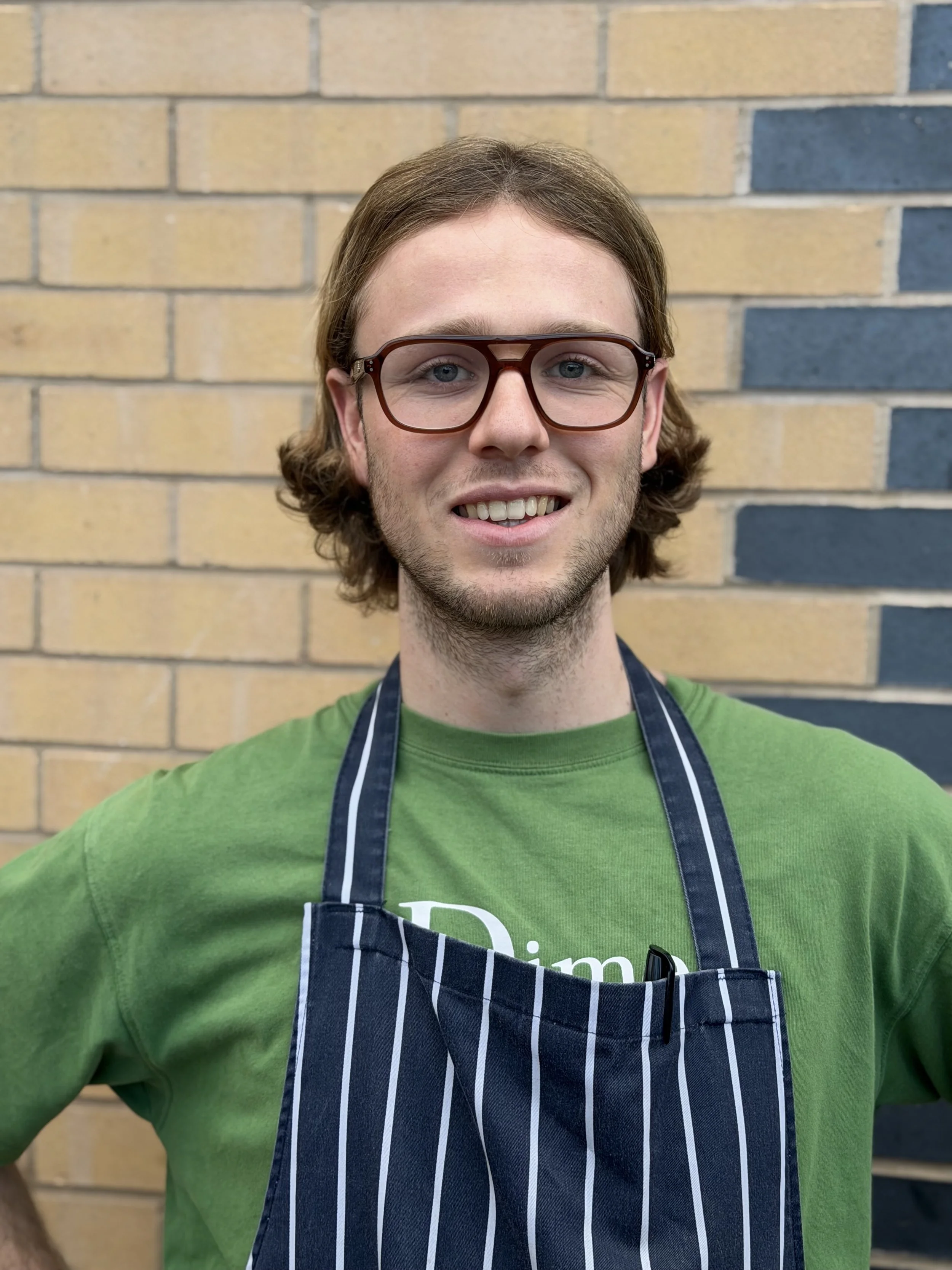 A young man with shoulder-length brown hair, wearing glasses, a green T-shirt, and a black and white striped apron, standing against a brick wall background.