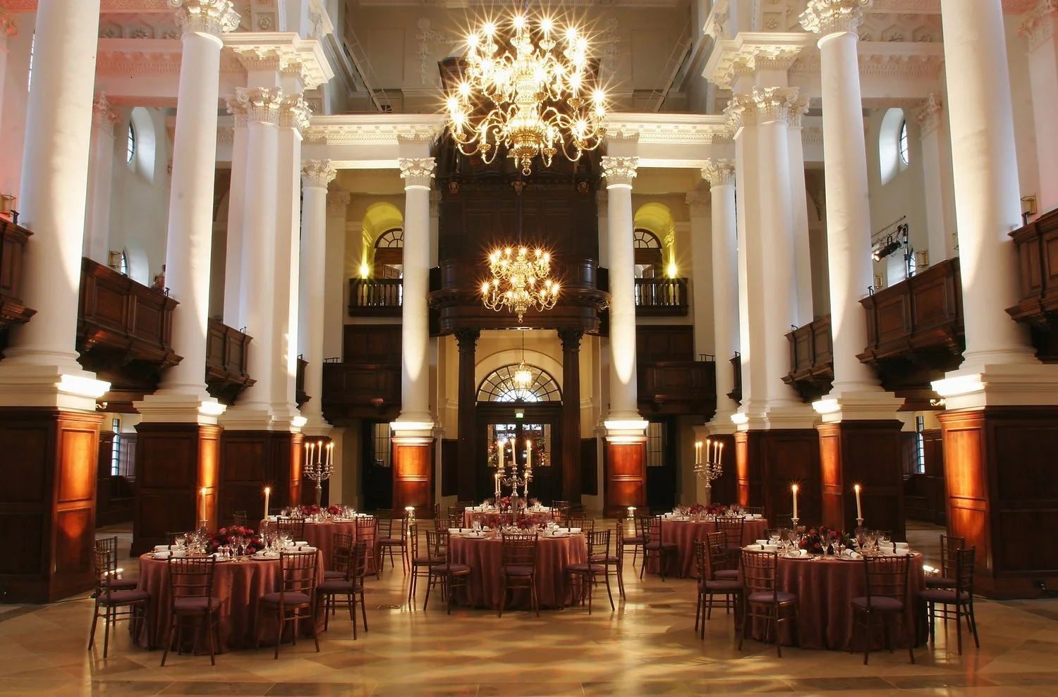 Elegant banquet hall with high ceilings, large white columns, wooden paneling, and chandeliers. Round tables with burgundy tablecloths, fine glassware, and floral centerpieces. Candles on tables and dim lighting create a sophisticated ambiance.